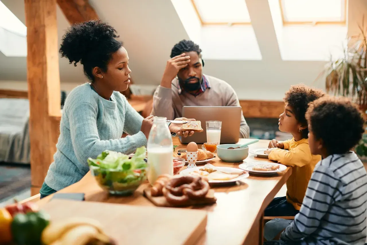 Family at a table with breakfast; father with head in hand, mother talking, children listening. Sunlight.