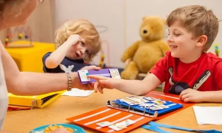 A child in a red shirt points at a game, another child looks on, a person assists. Teddy bear in background.