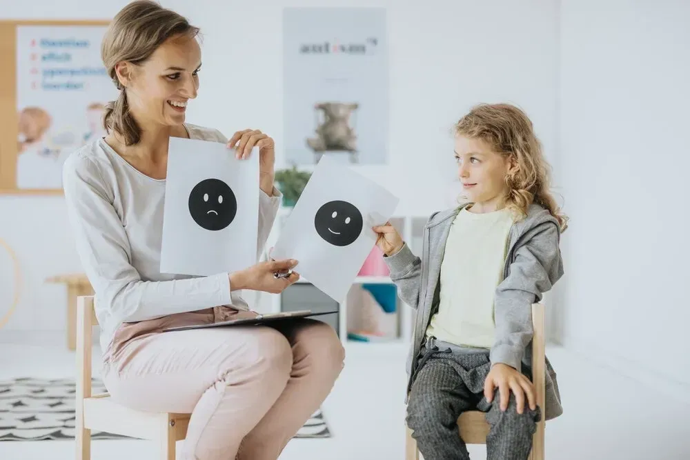 Woman holding sad face illustrations, interacting with child in therapy setting.
