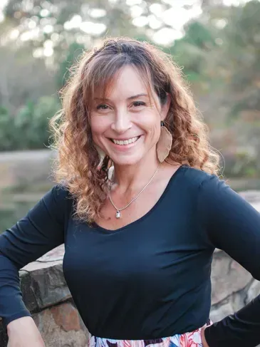 Woman with curly brown hair smiles, wearing a black top, earrings, and necklace, outdoors.