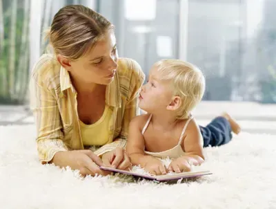 Woman reading to a toddler lying on a white rug; both looking at a book.