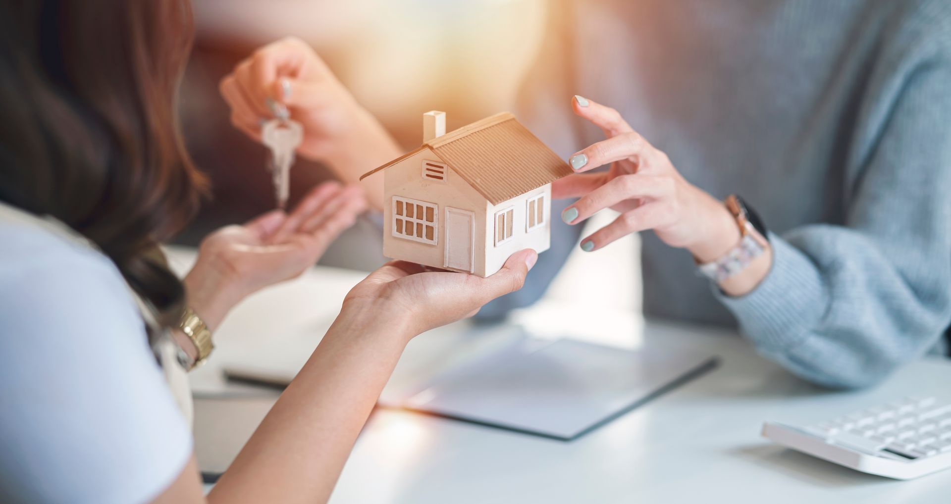 Person handing keys and a miniature house to another person at a table.