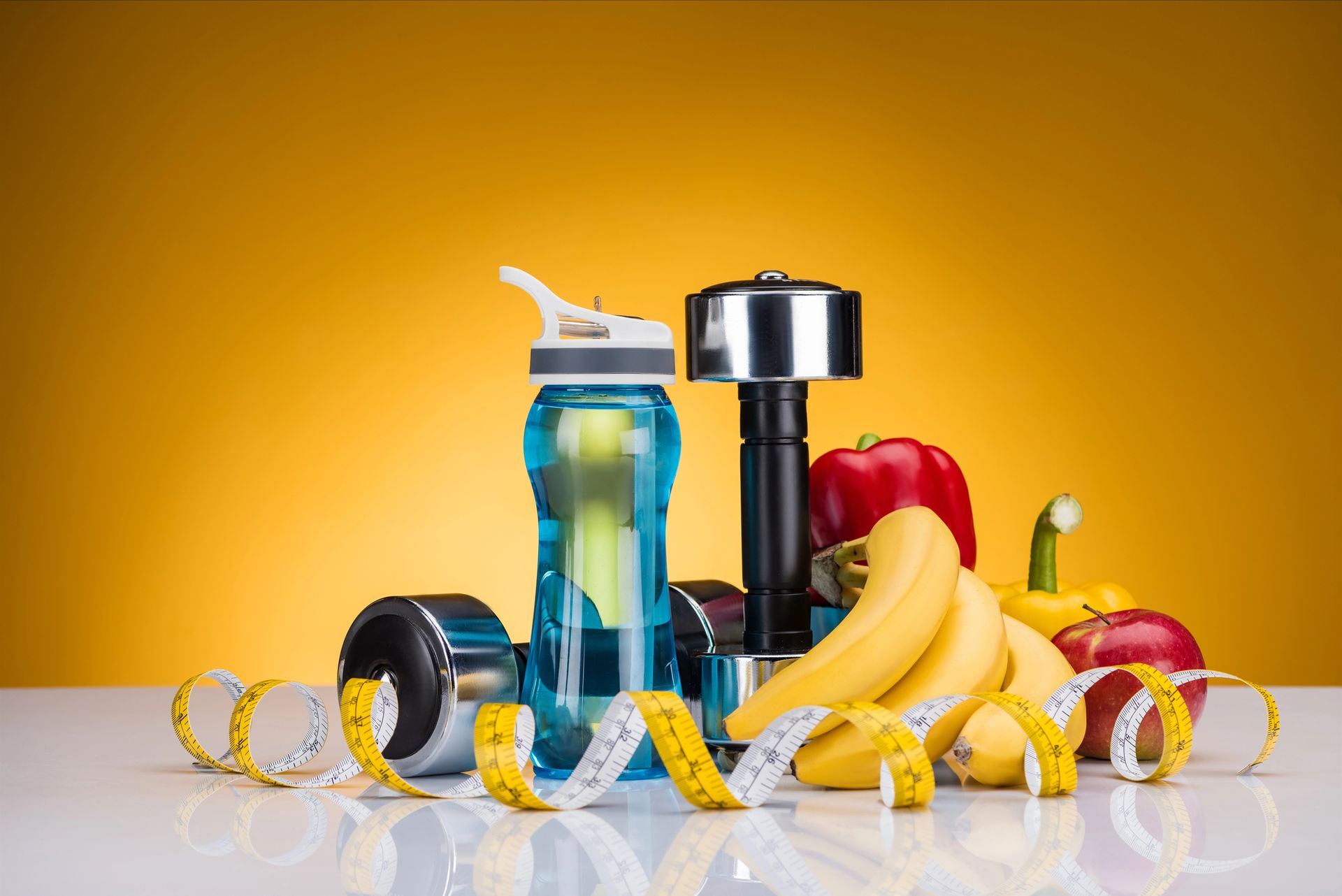 Equipo de fitness con botella de agua, mancuerna, plátanos, manzanas y cinta métrica sobre un fondo amarillo.