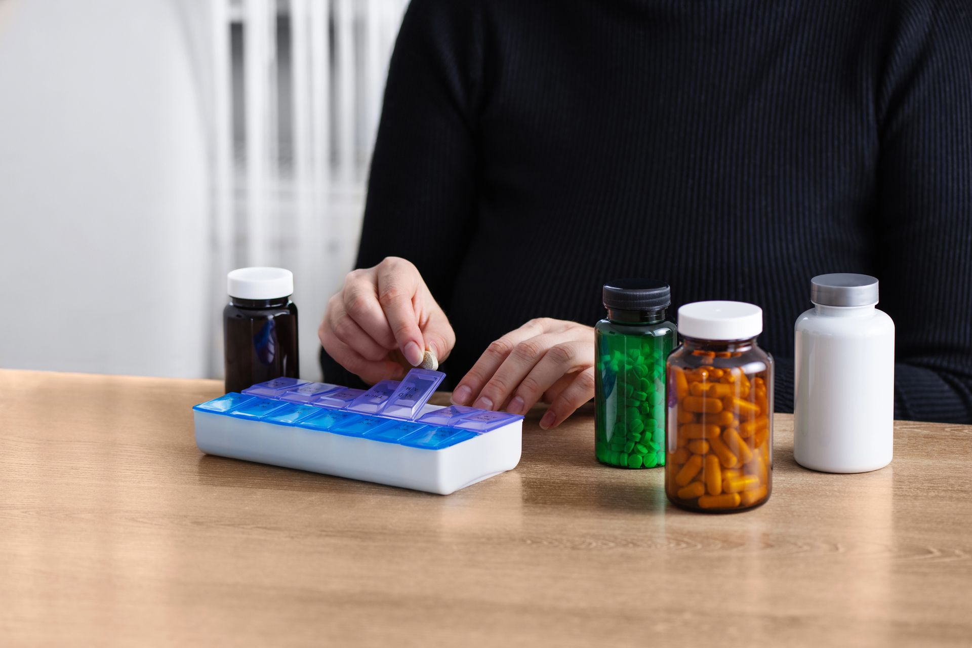 Manos colocando una pastilla en un organizador semanal junto a frascos de medicamentos sobre una mesa.