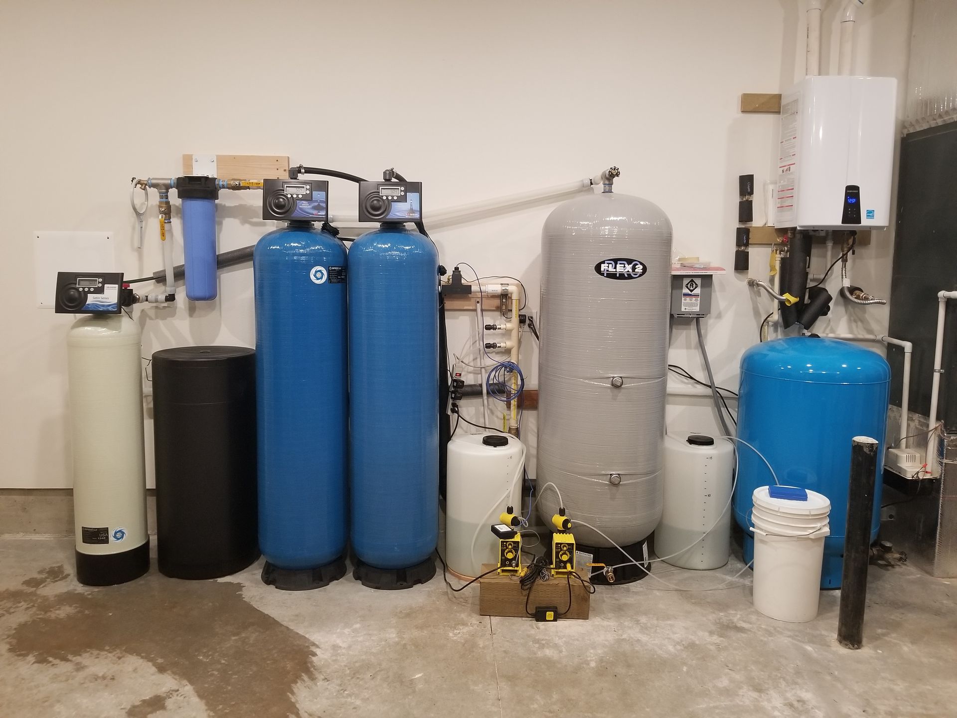 Row of water treatment tanks and filters in a utility room, mostly blue, gray, and black.