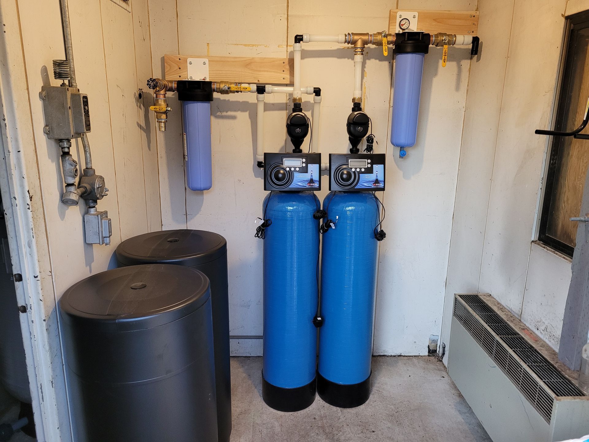 Water treatment room with blue filter tanks, black brine tanks, and wall-mounted plumbing.