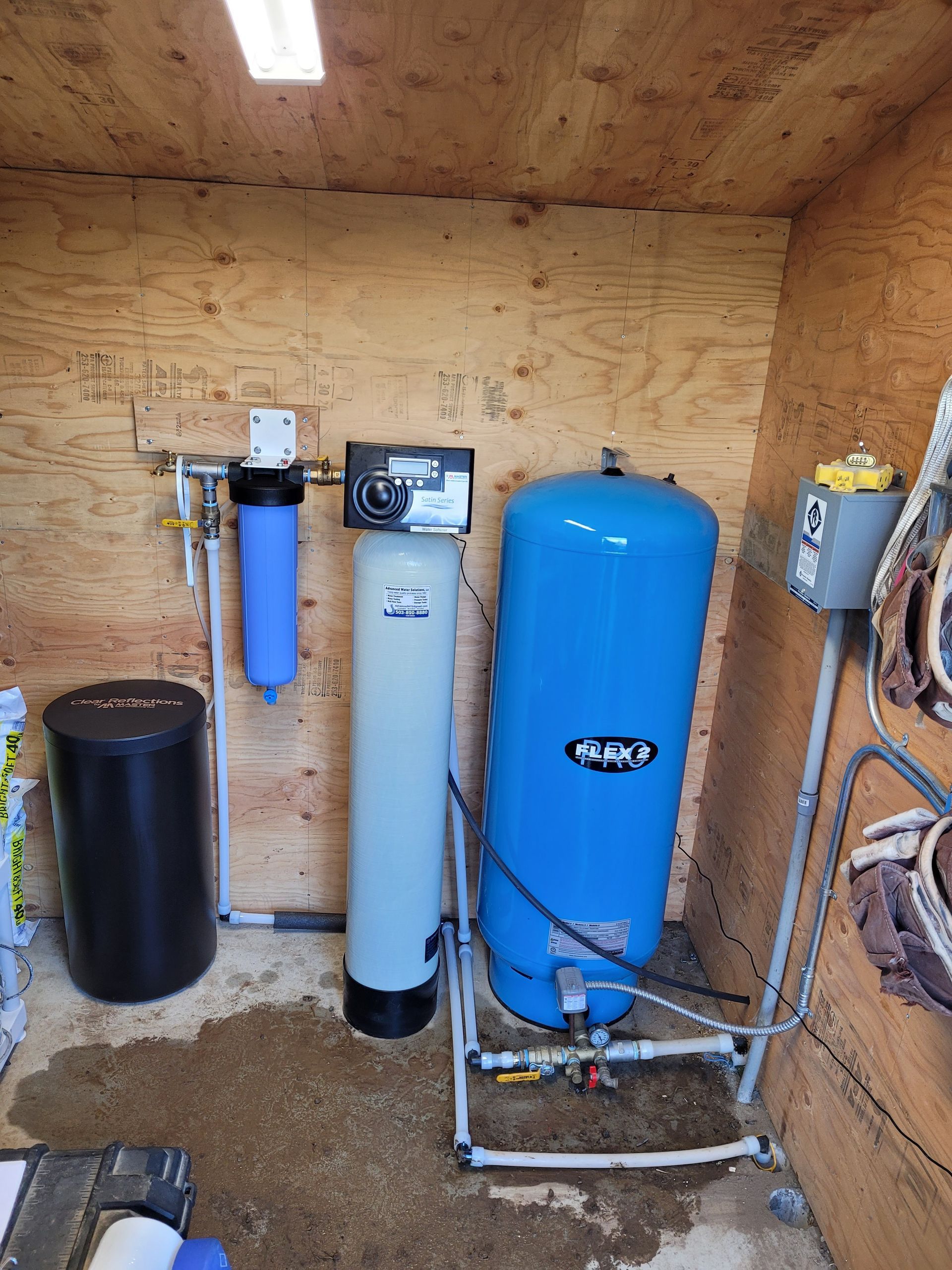 Water treatment equipment in a wooden utility room, with blue and white filtration tanks and pipes.