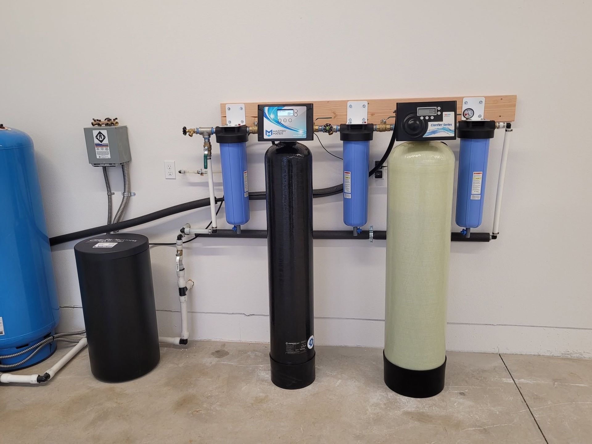 Wall-mounted water filtration system with several large blue, black, and white filter tanks in a utility room