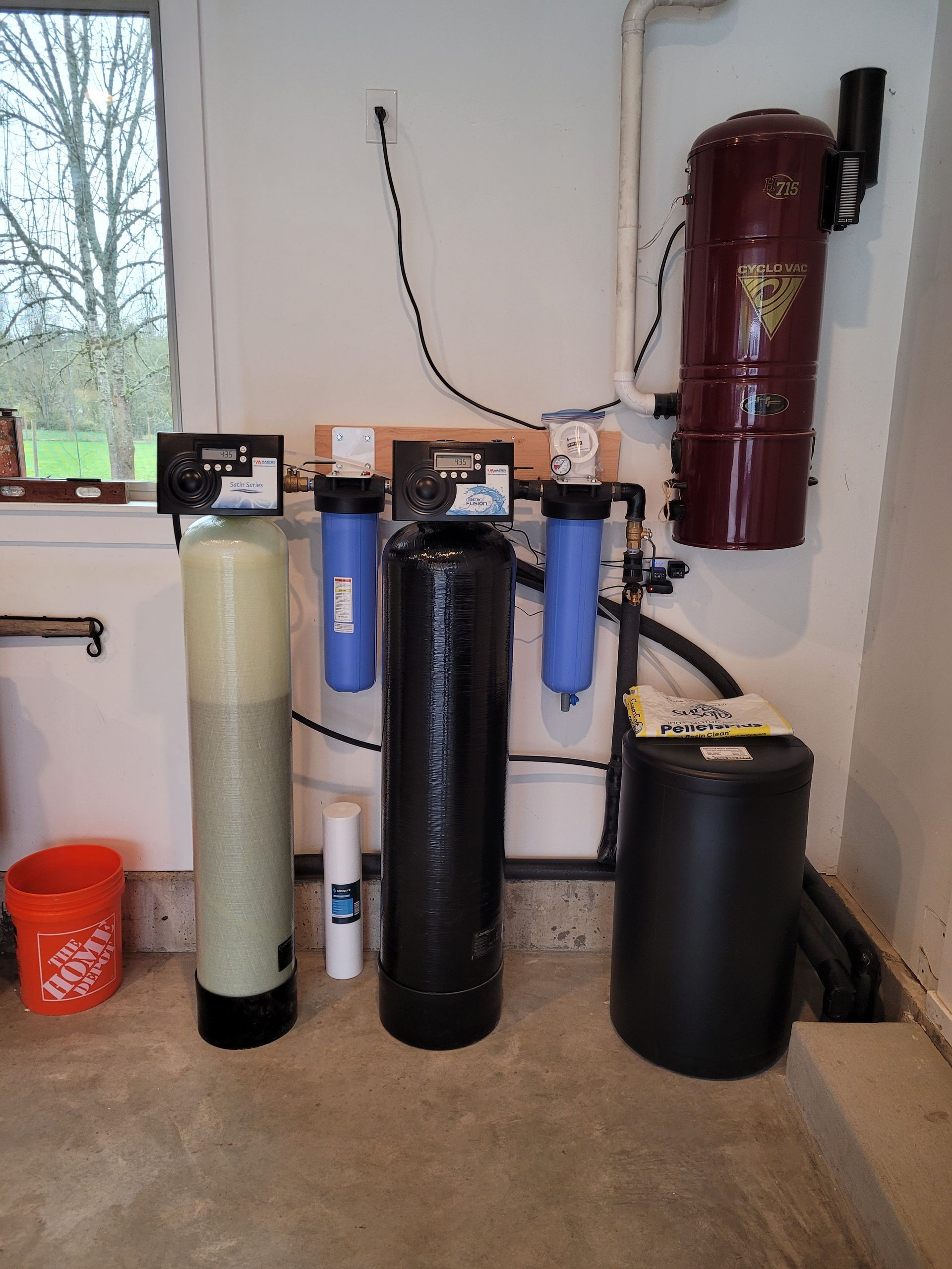 Water softener and filtration tanks in a utility room beside a window and red wall-mounted cylinder.