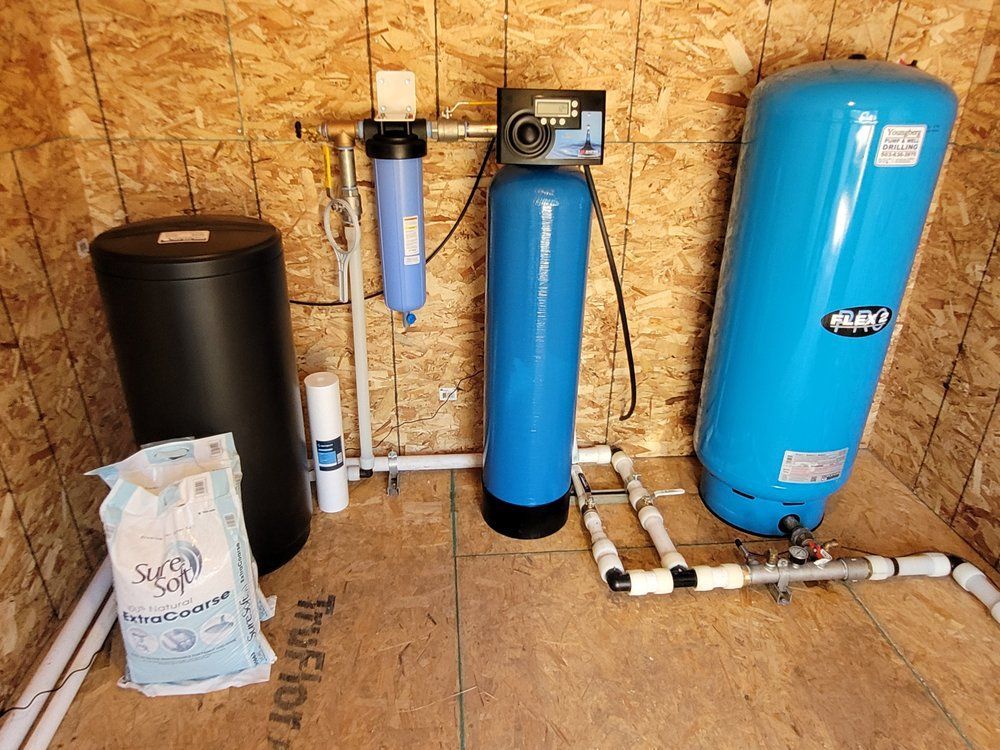 Water softener and filtration system in a utility room, with blue and black tanks and white plumbing pipes.