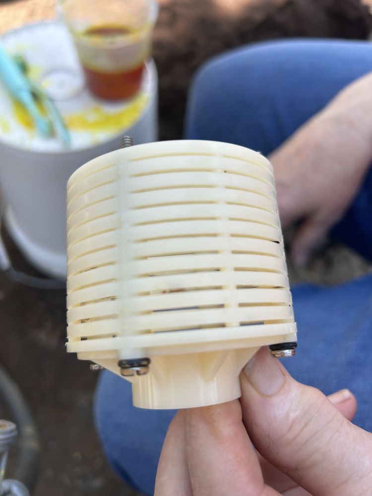 Hand holding a small white vented cylindrical plastic part outdoors, with a blurred drink and leg in background