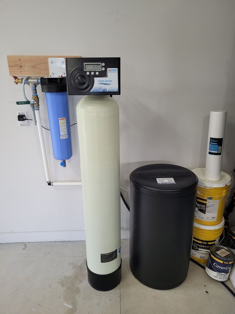 Water softener and filtration system installed against a garage wall, with black and blue tanks and filter canisters.