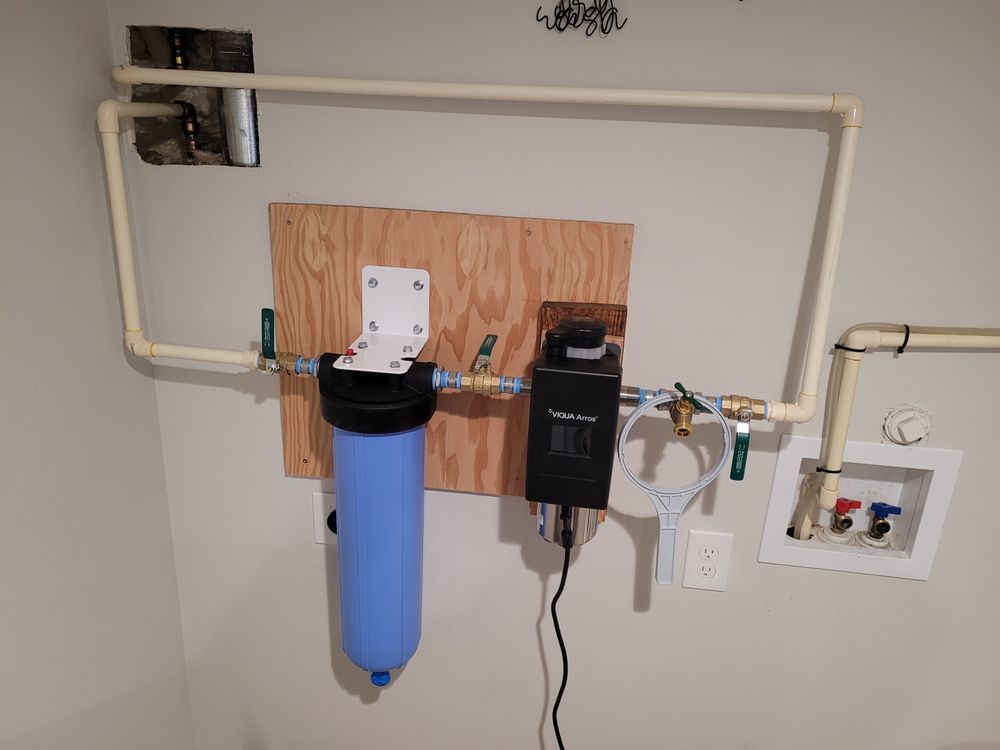 Wall-mounted water filtration system with blue filter canister, white pipes, and control unit in a utility room