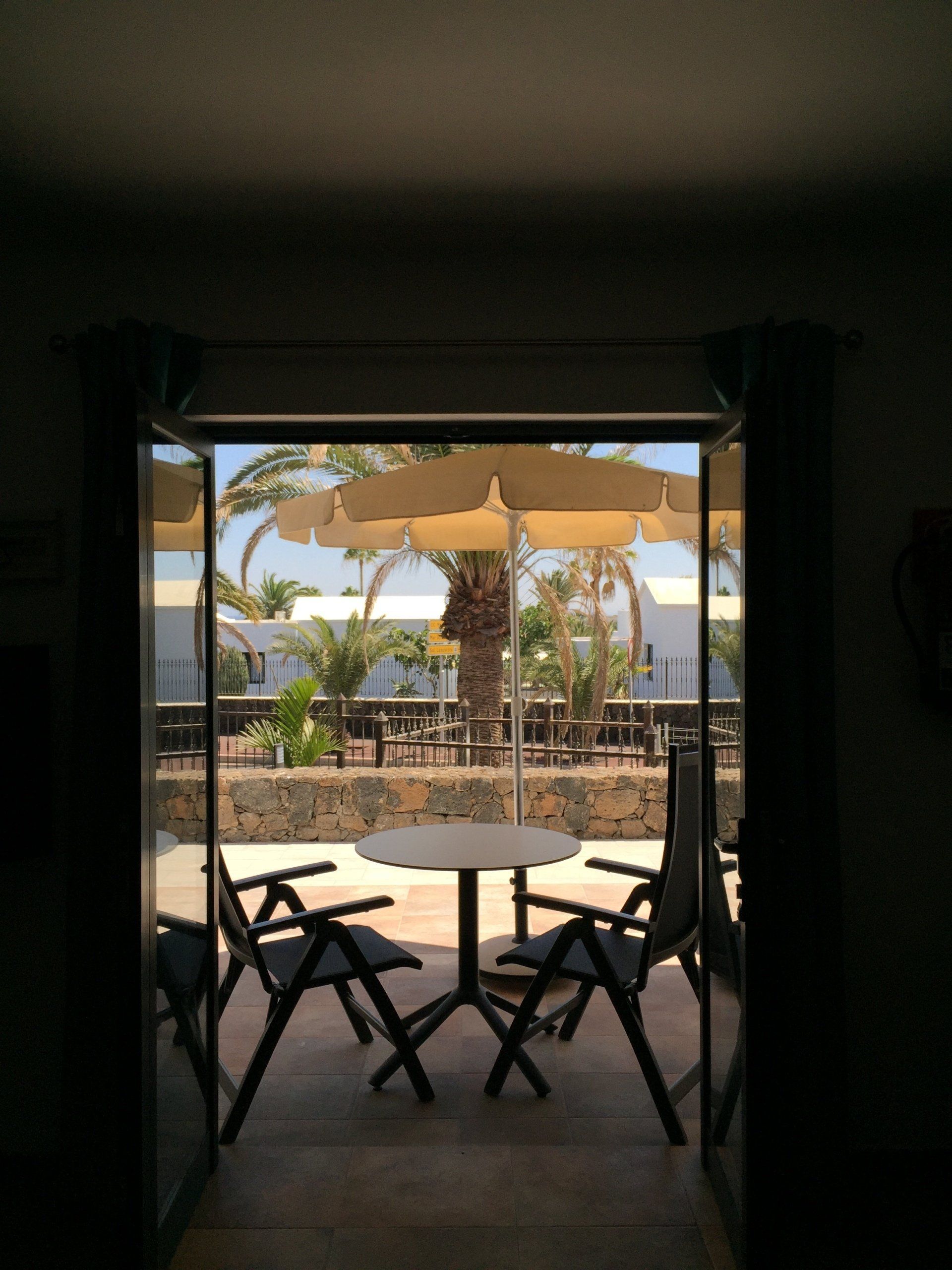 Vista a través de una puerta abierta a un patio con una mesa, sillas y una sombrilla, en un entorno soleado.