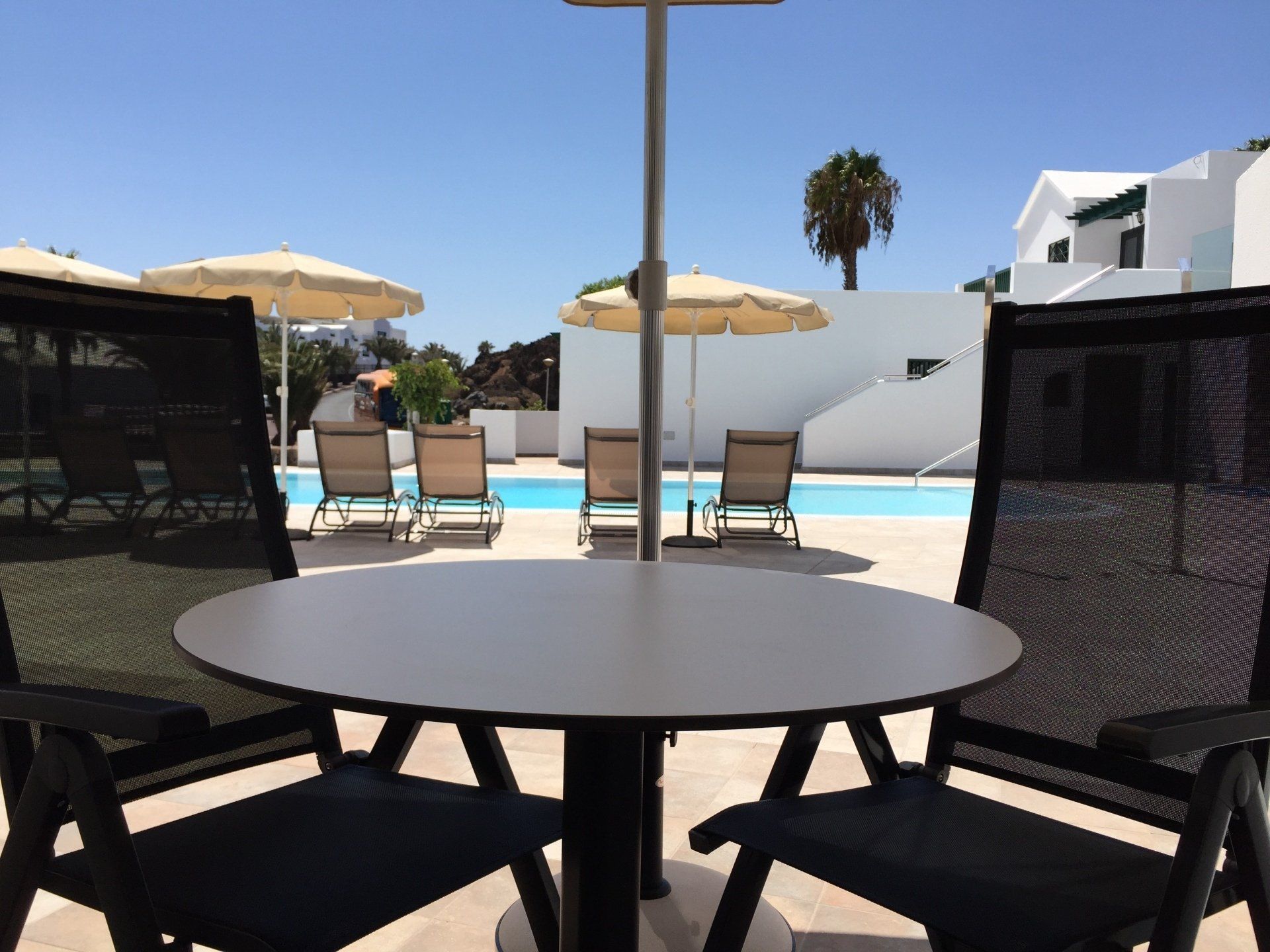 Una escena de piscina soleada: mesa y sillas en primer plano, tumbonas y sombrillas junto a la piscina, edificios blancos.