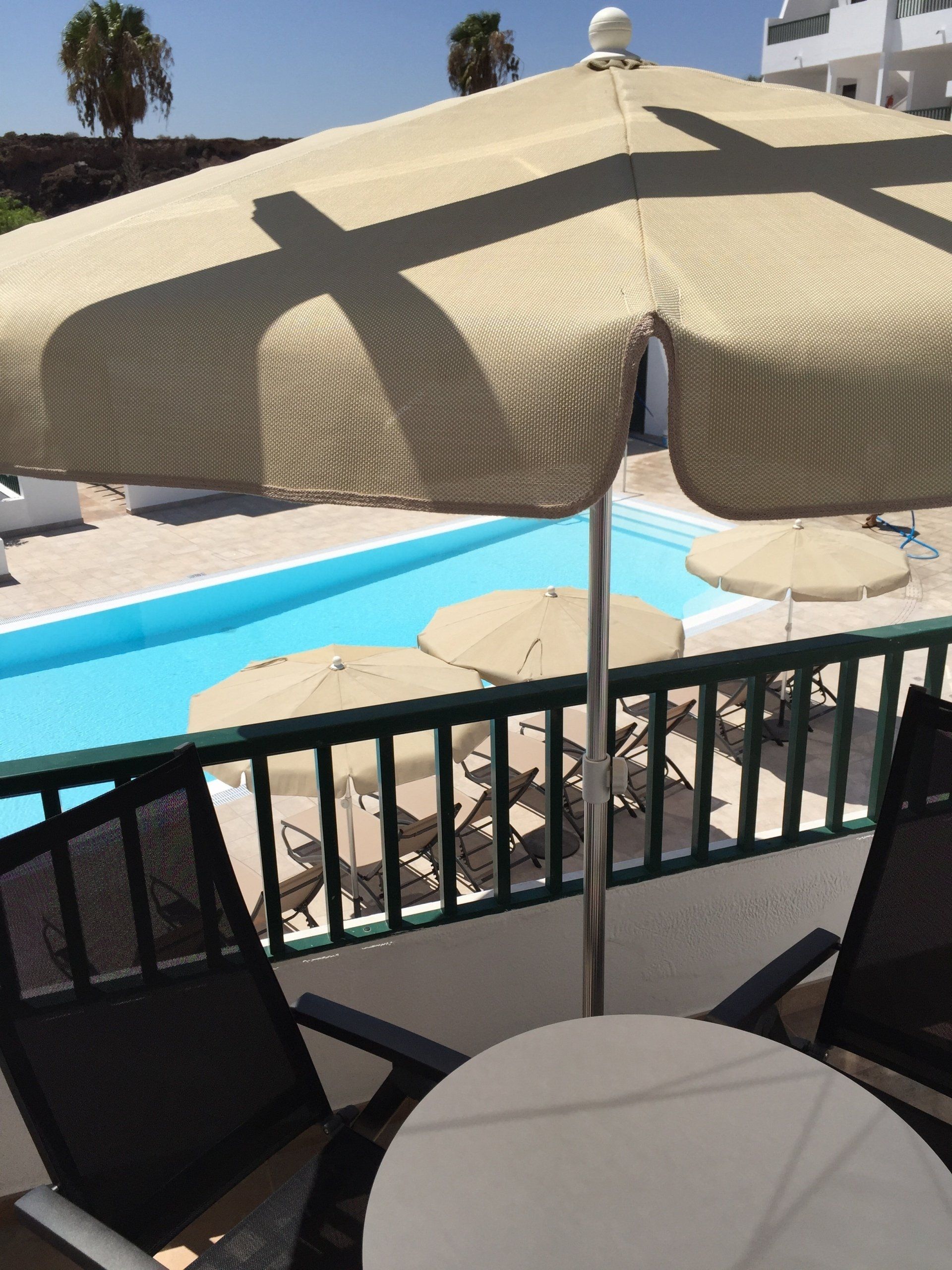 Patio con mesa, sillas y una gran sombrilla beige con vista a una piscina con otras sombrillas.