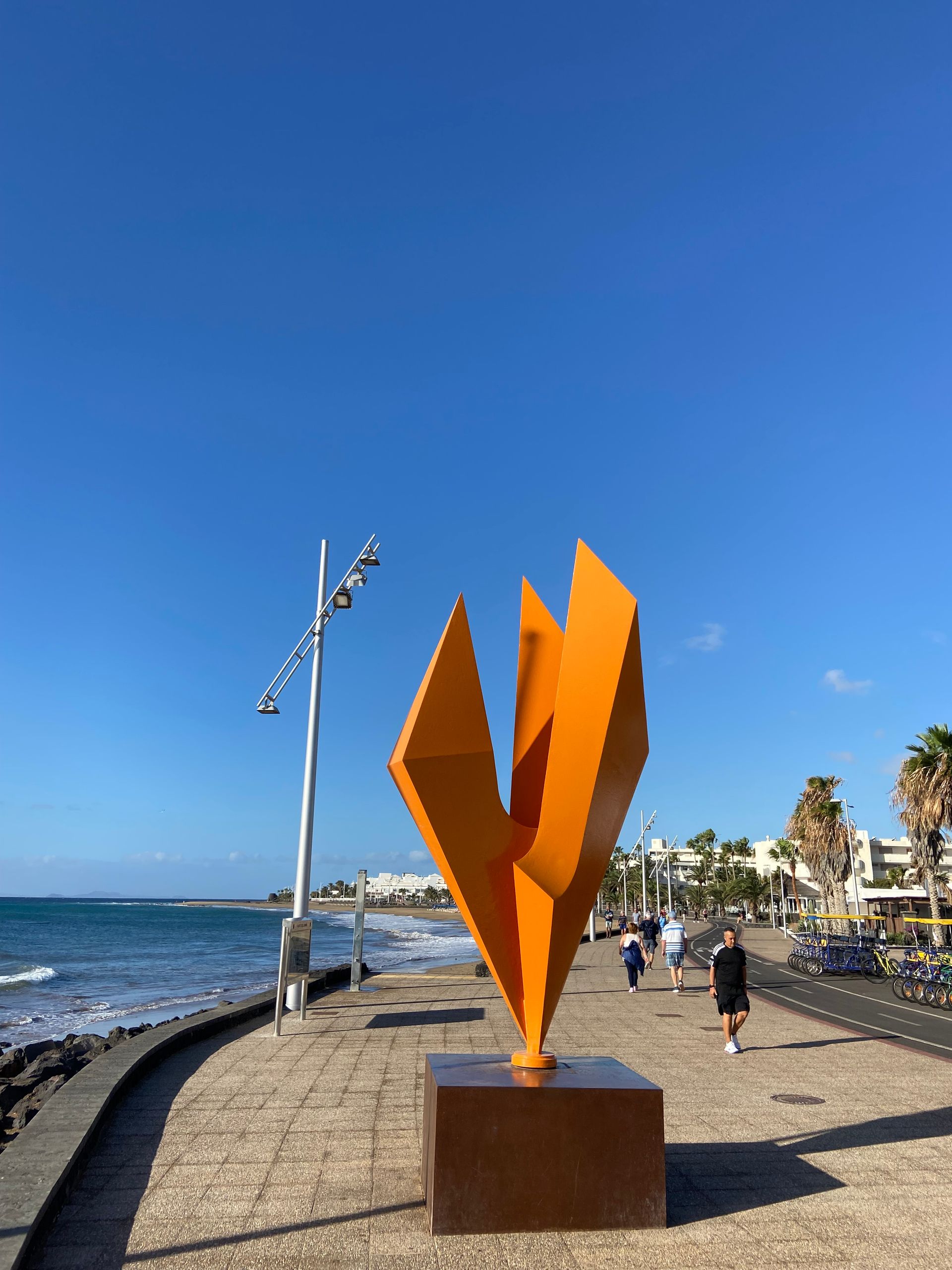 Orange abstract sculpture on a seaside promenade, blue sky, people walking, palm trees.