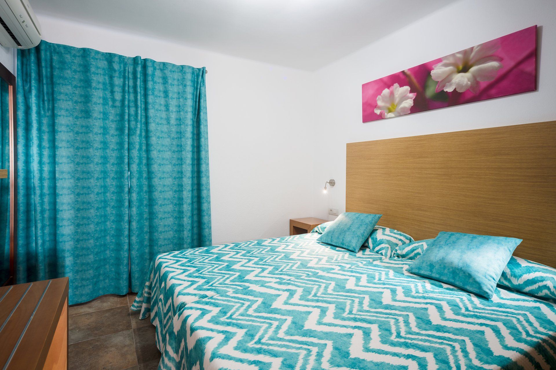 Dormitorio con cortinas, colcha y almohadas color verde azulado. Cabecero de madera, obra de arte y mesita de noche.