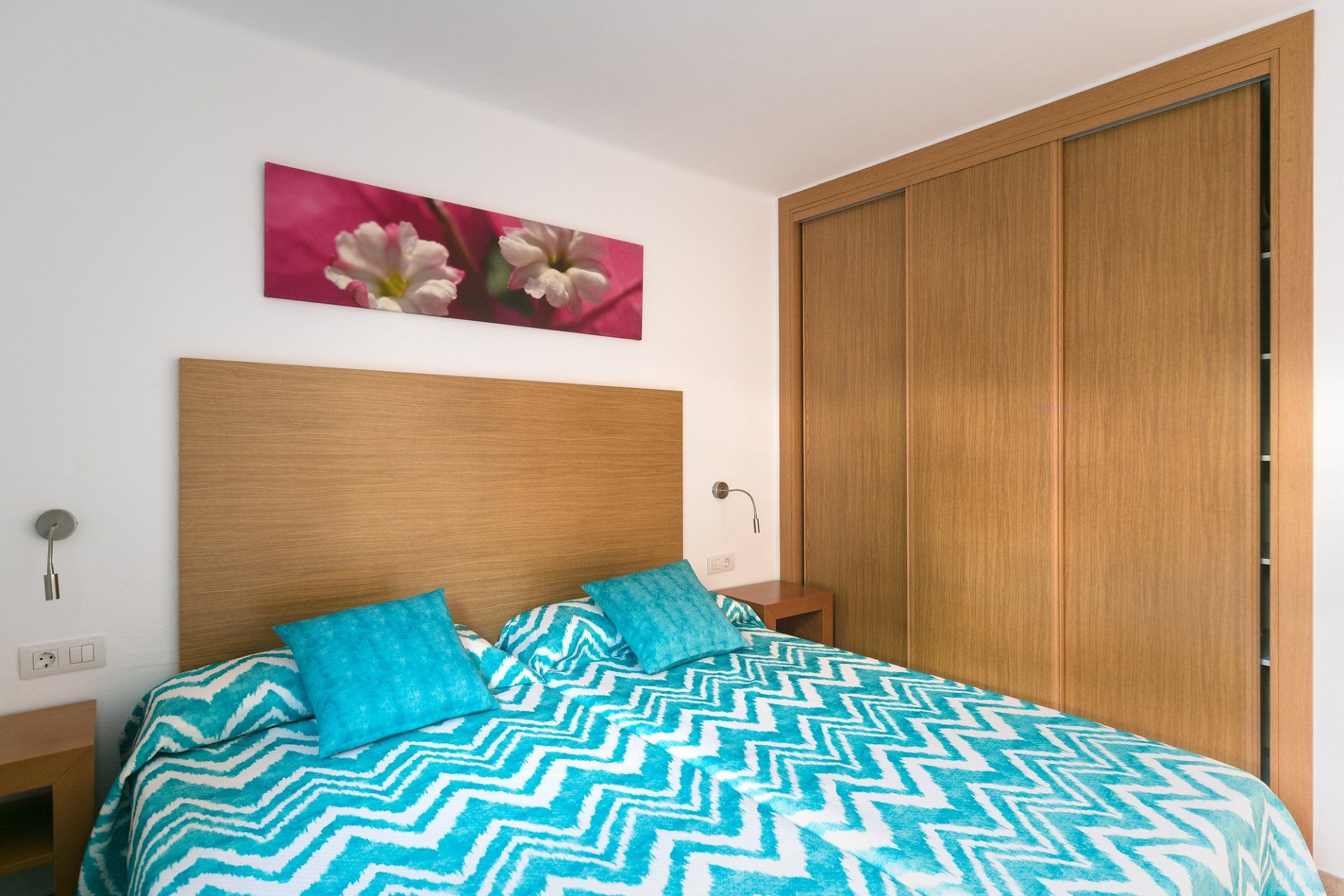 Dormitorio con cama con ropa de cama estampada en turquesa y blanco, cabecero de madera, armario y arte floral.