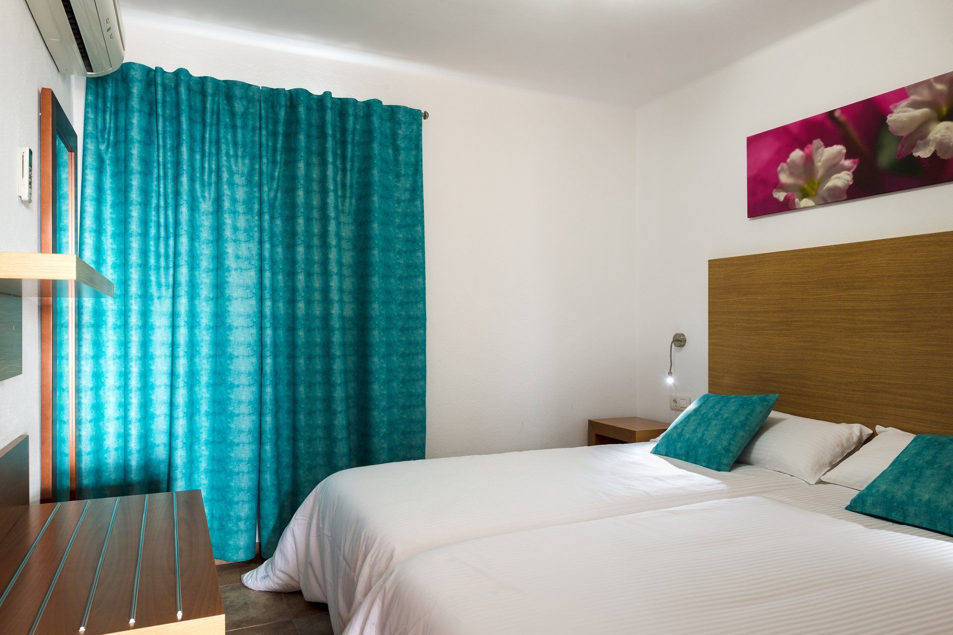Dormitorio con dos camas individuales, cortinas color verde azulado, paredes blancas y obras de arte florales.
