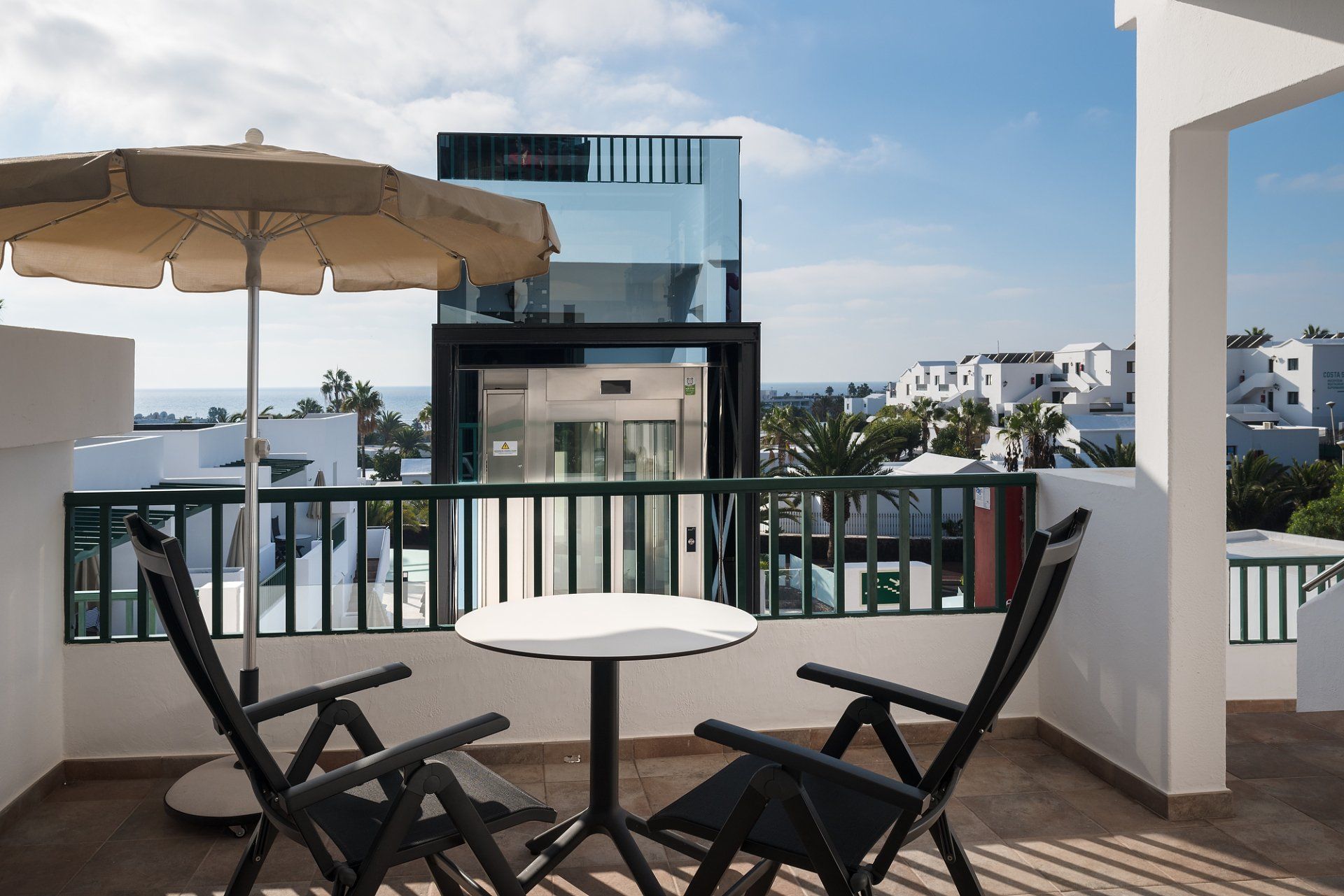 Balcón con mesa y sillas, vista al mar. Un moderno edificio de cristal al fondo. Día soleado.