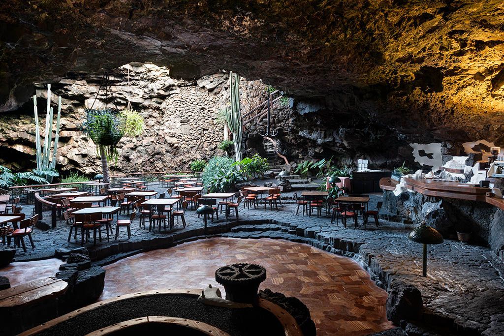 Restaurant inside a lava cave with tables, chairs, and vegetation, lit by warm lights.