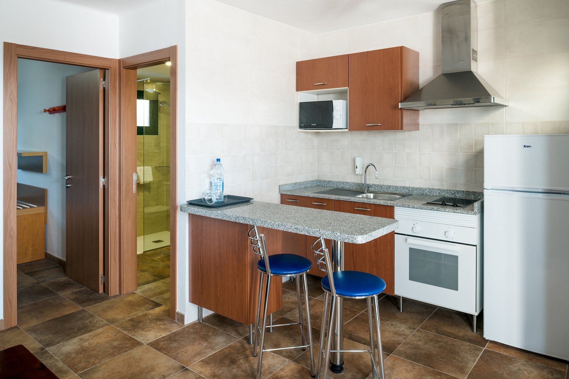 Cocina pequeña con armarios de madera, electrodomésticos blancos, una barra de desayuno y dos taburetes azules.