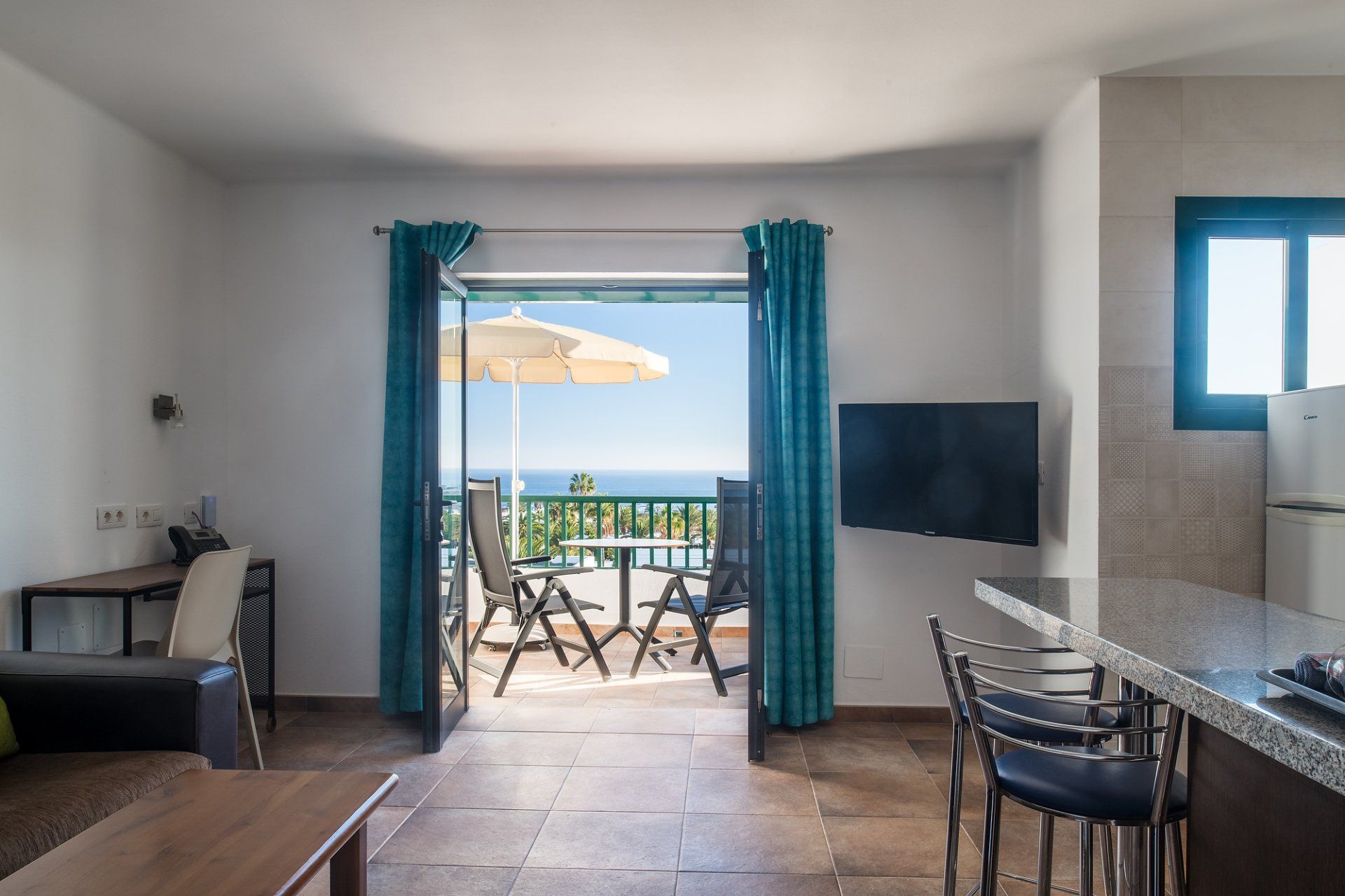 Vista interior: Espacio de estar con balcón abierto con vista al mar; cortinas verde azulado, mesa/sillas de exterior.
