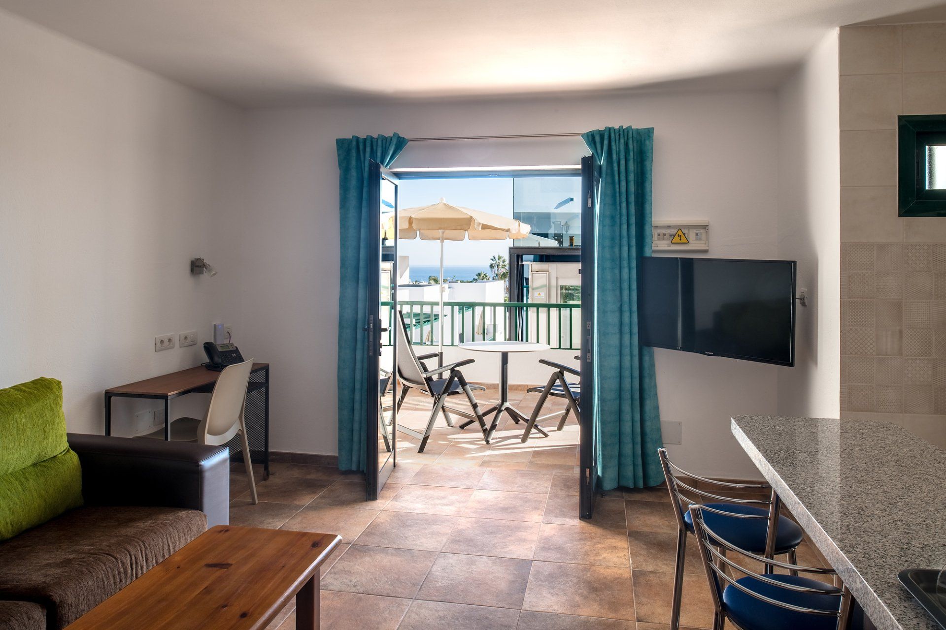 Salón moderno con sofá, escritorio, televisor y puertas que dan a un patio con sillas y vistas al mar.