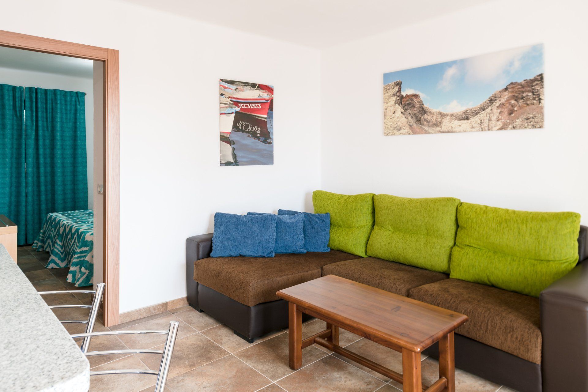 Sala de estar con sofá marrón, cojines verdes y azules, mesa de madera, arte y una vista de un dormitorio.