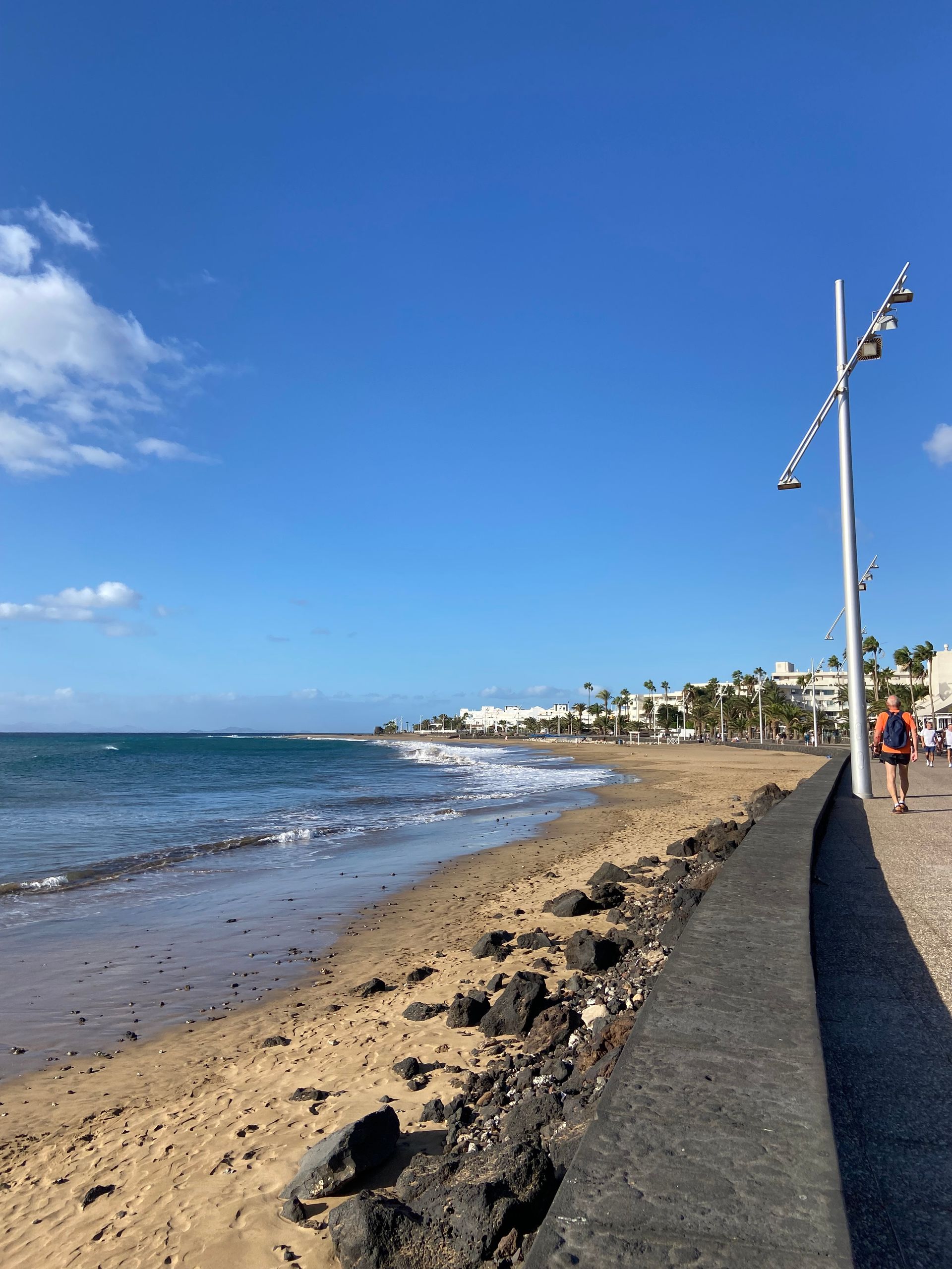 Beach scene with sandy shore, blue water, and sky. A person walks on a path.
