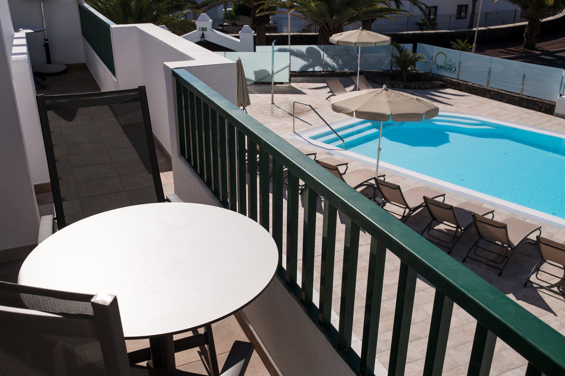 Balcón con mesa y sillas con vista a una piscina con reposeras y sombrillas.