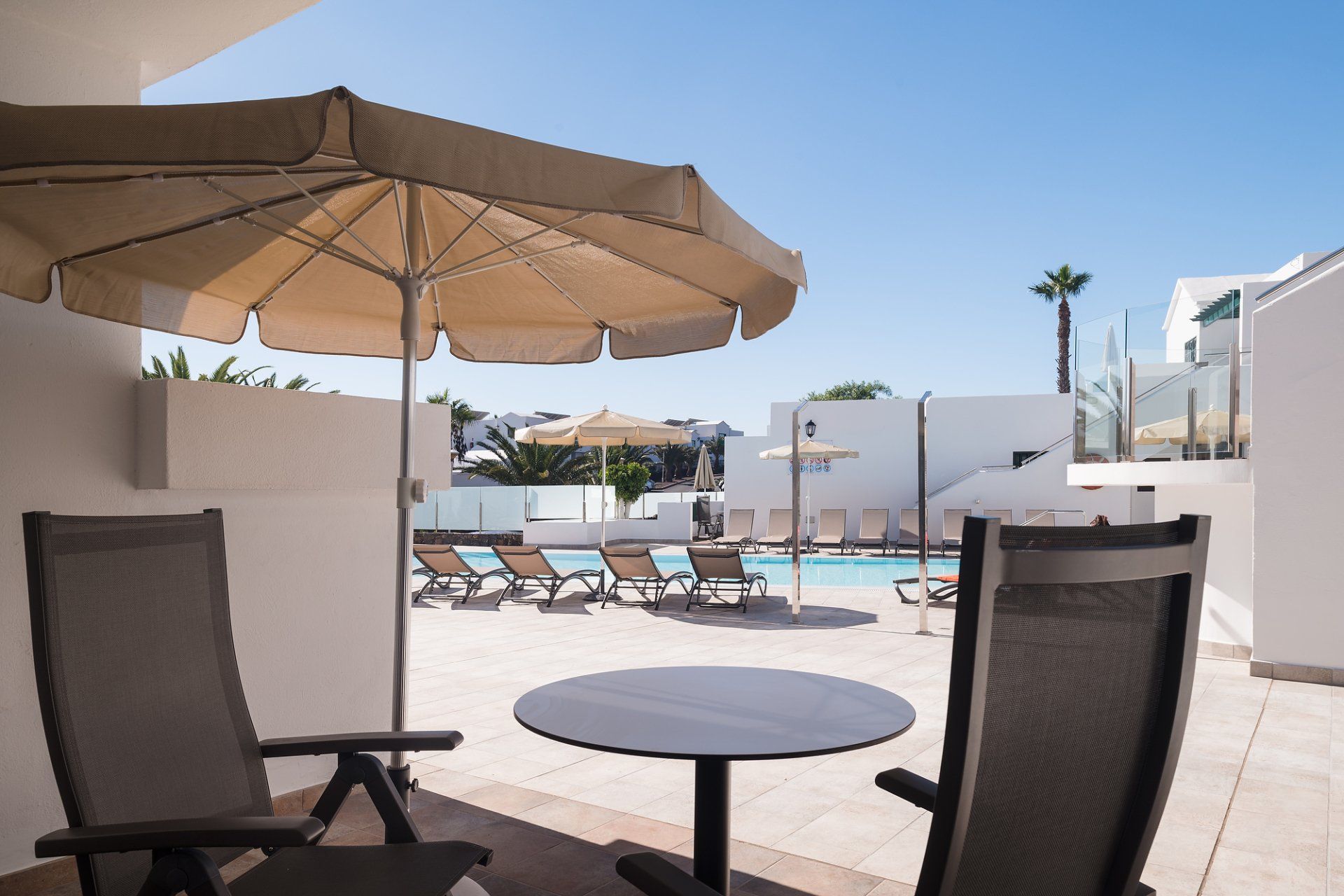Patio con sillas, mesa y sombrilla con vista a un área de piscina con reposeras en un lugar soleado.