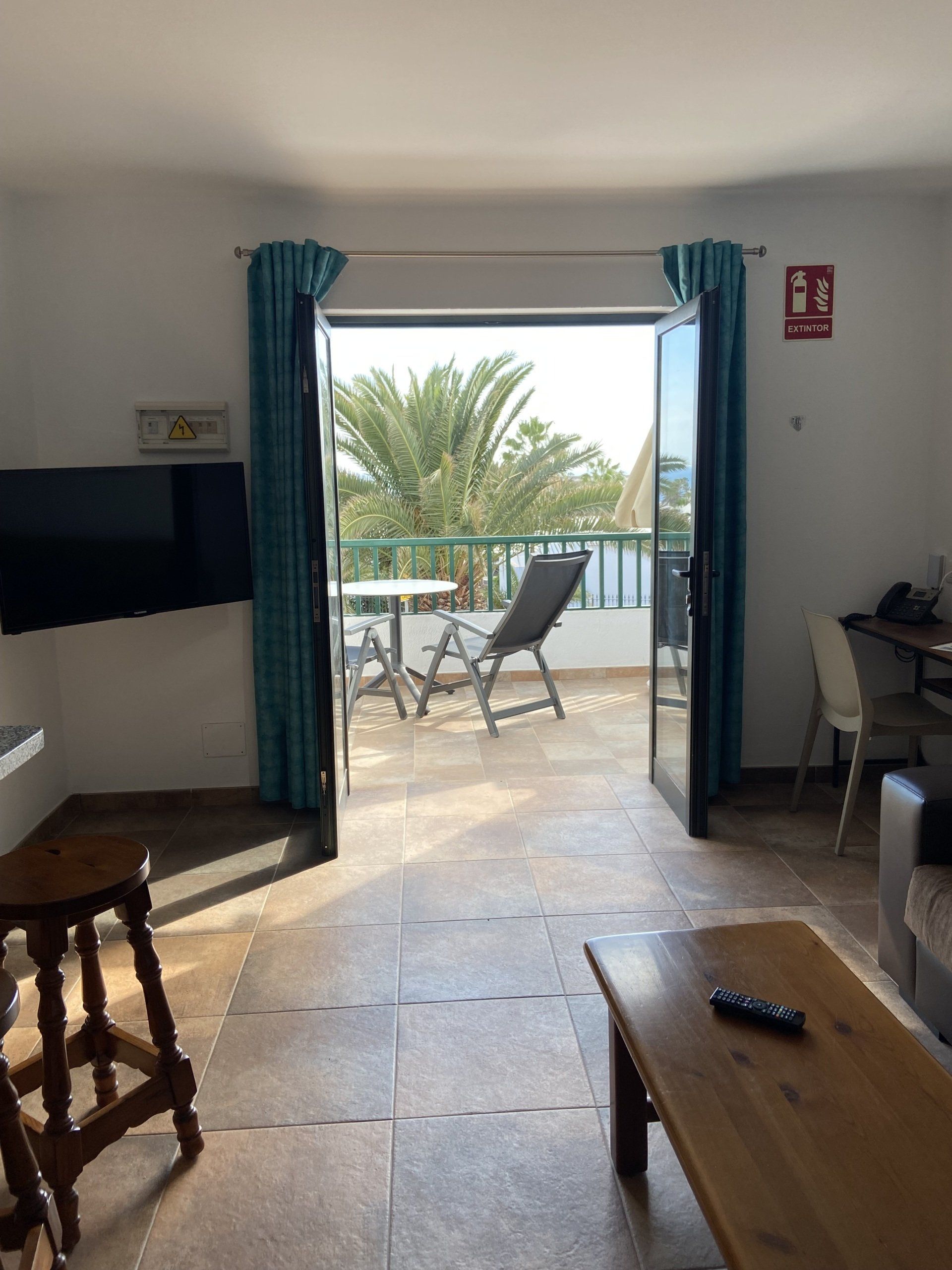 Interior con puertas abiertas a un balcón; cortinas turquesas, piso de baldosas y vista a las palmeras.