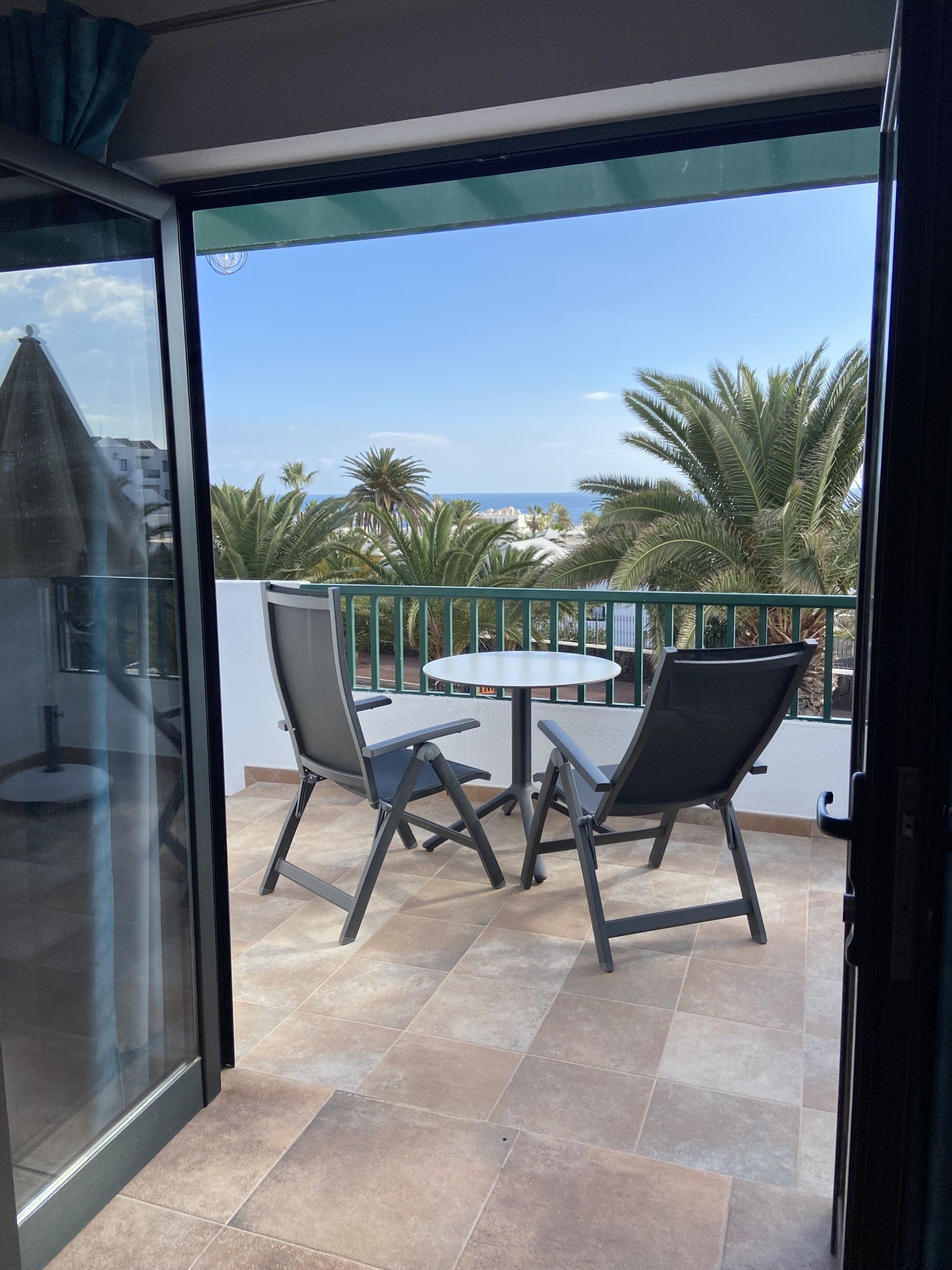 Balcón con sillas y mesa con vista a las palmeras y al mar bajo un cielo azul.