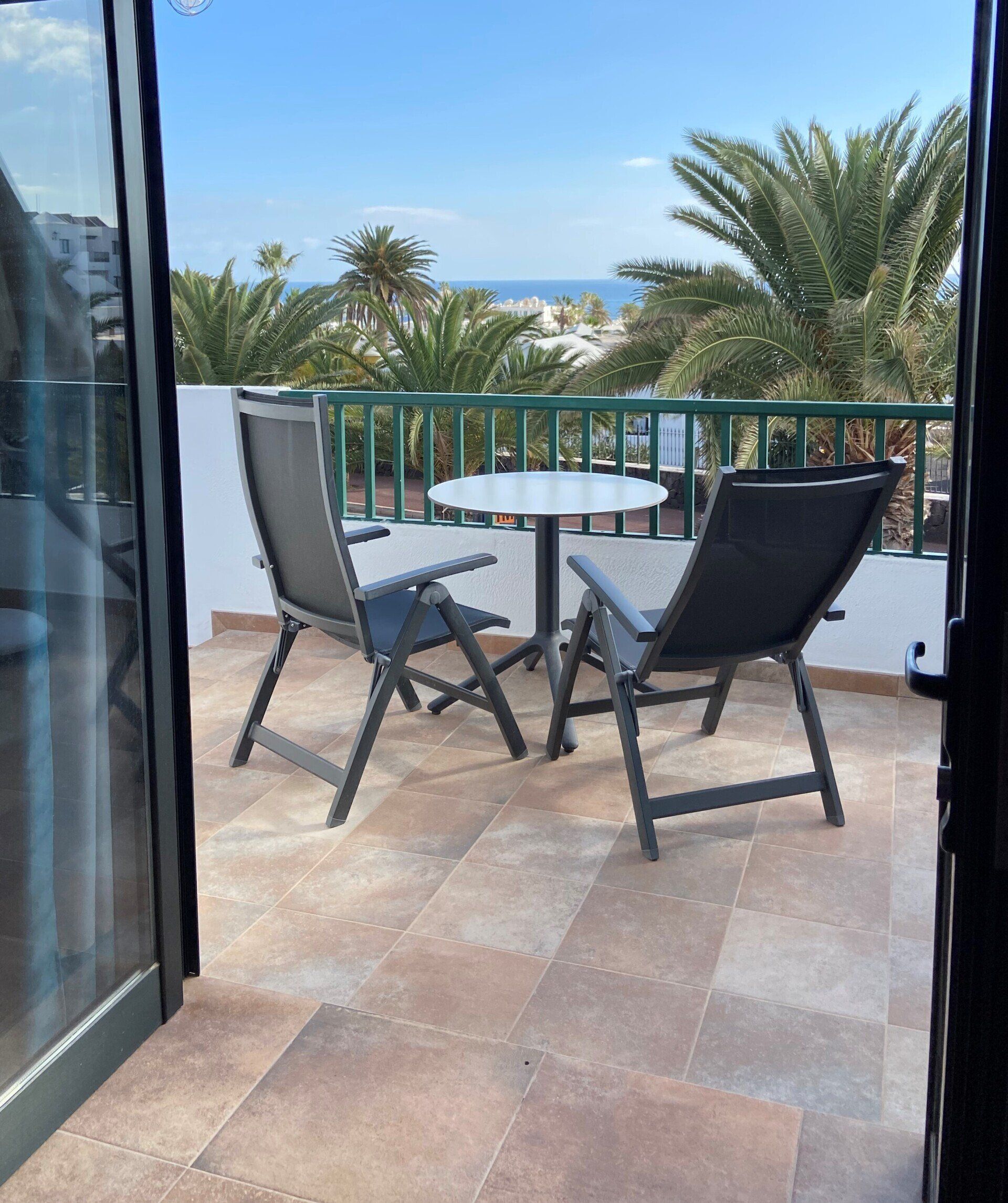 Balcón con dos sillas y mesa, con vista a un soleado mar con palmeras.