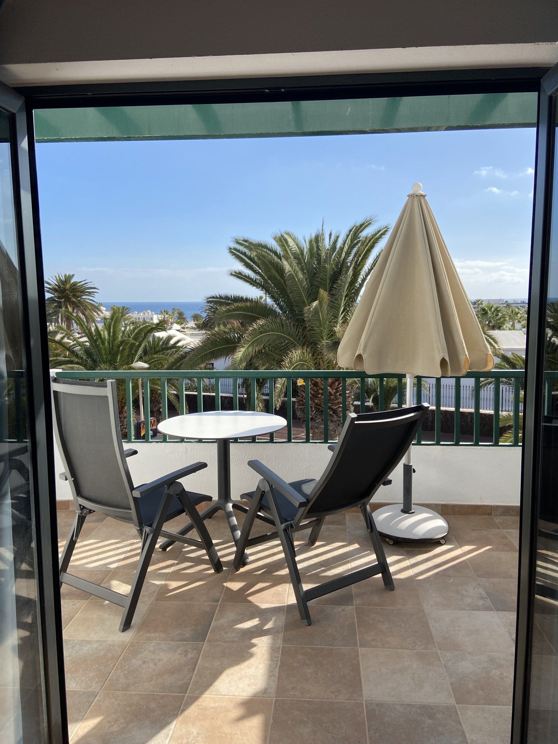 Vista desde el balcón: Dos sillas, una mesa y una sombrilla en un patio con vistas al mar. Palmeras y cielo azul.