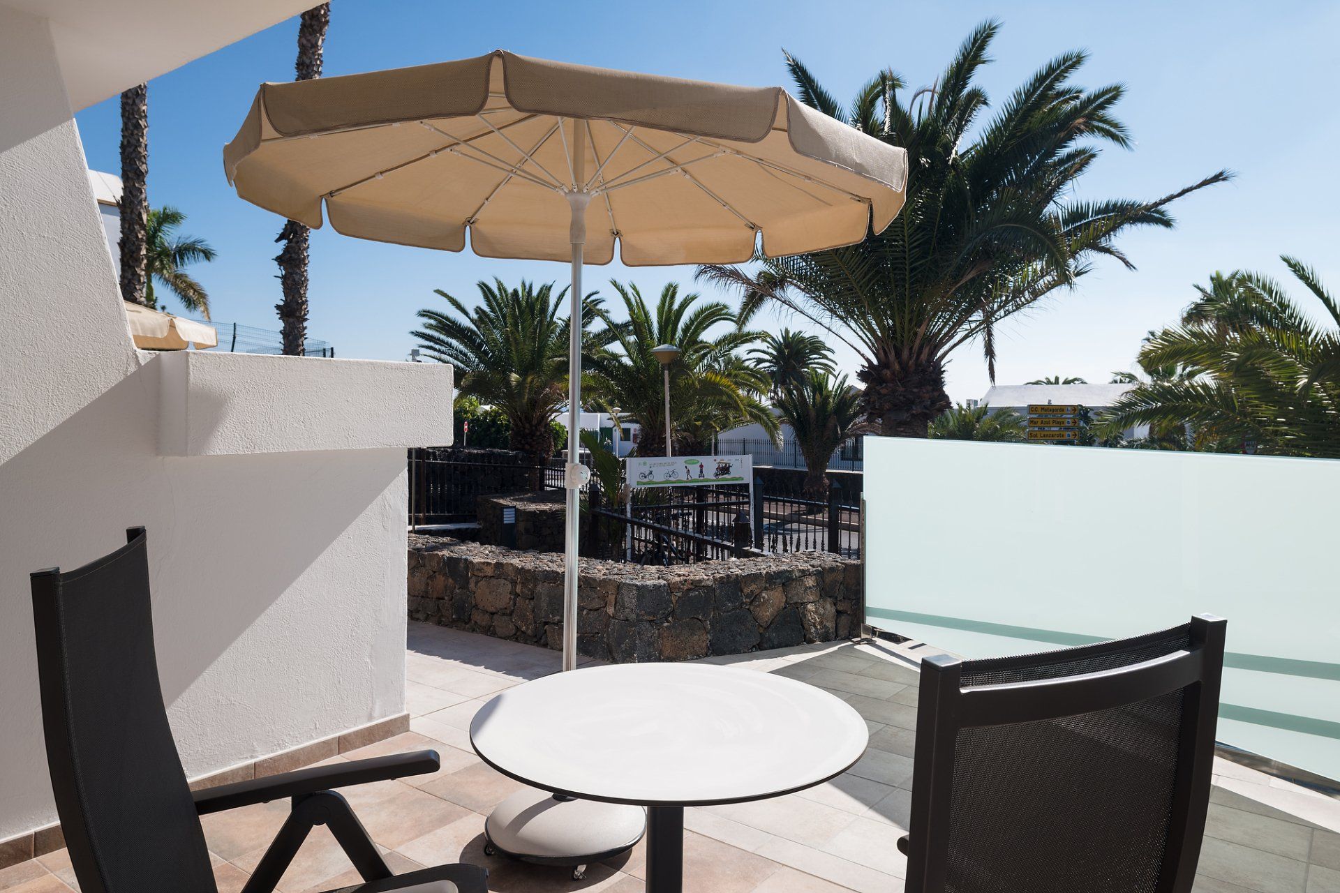 Balcón con mesa, sillas y sombrilla, con vista a un soleado comedor al aire libre.