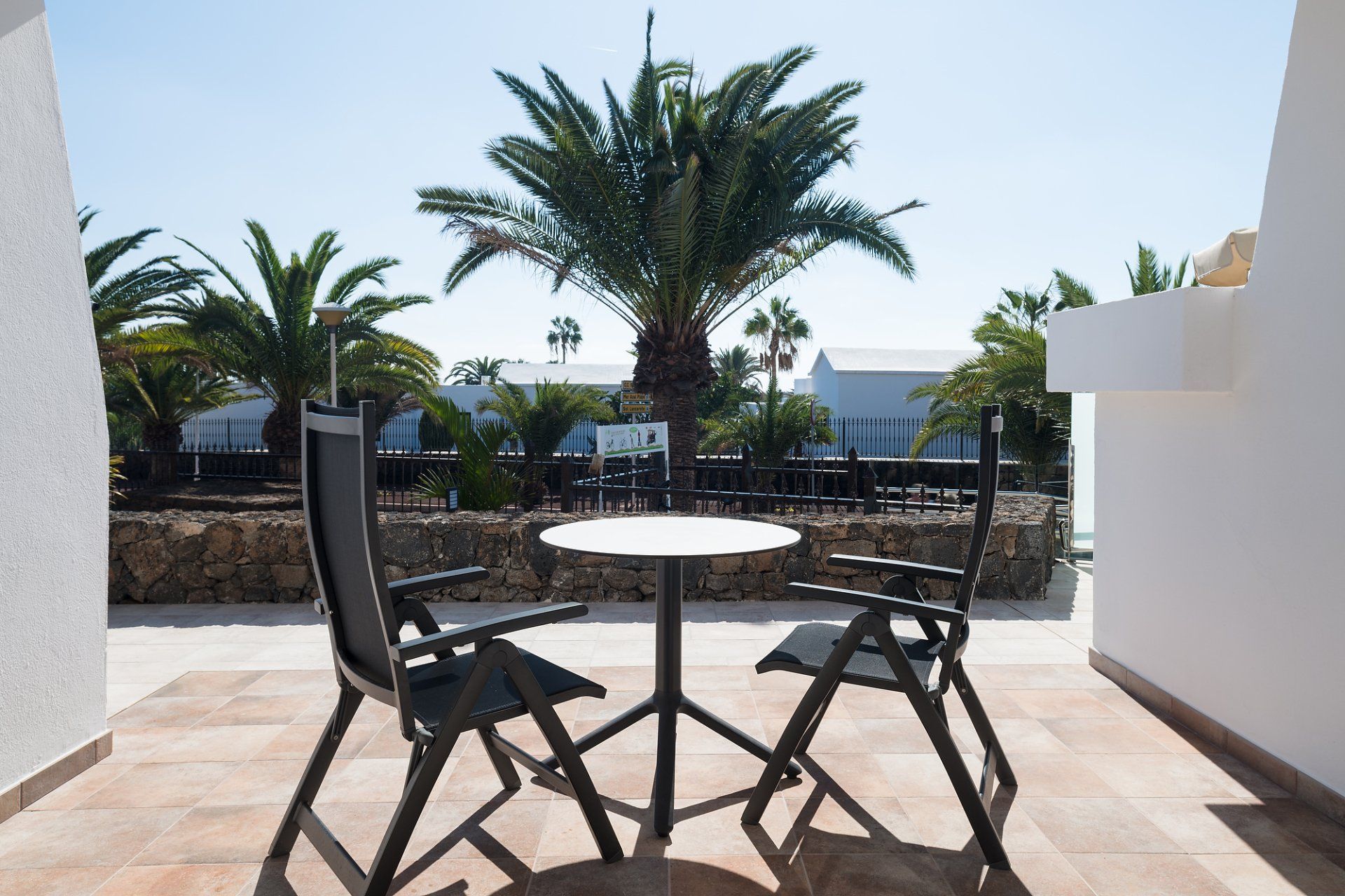 Patio con mesa, sillas, palmeras y edificios blancos bajo un cielo azul brillante.