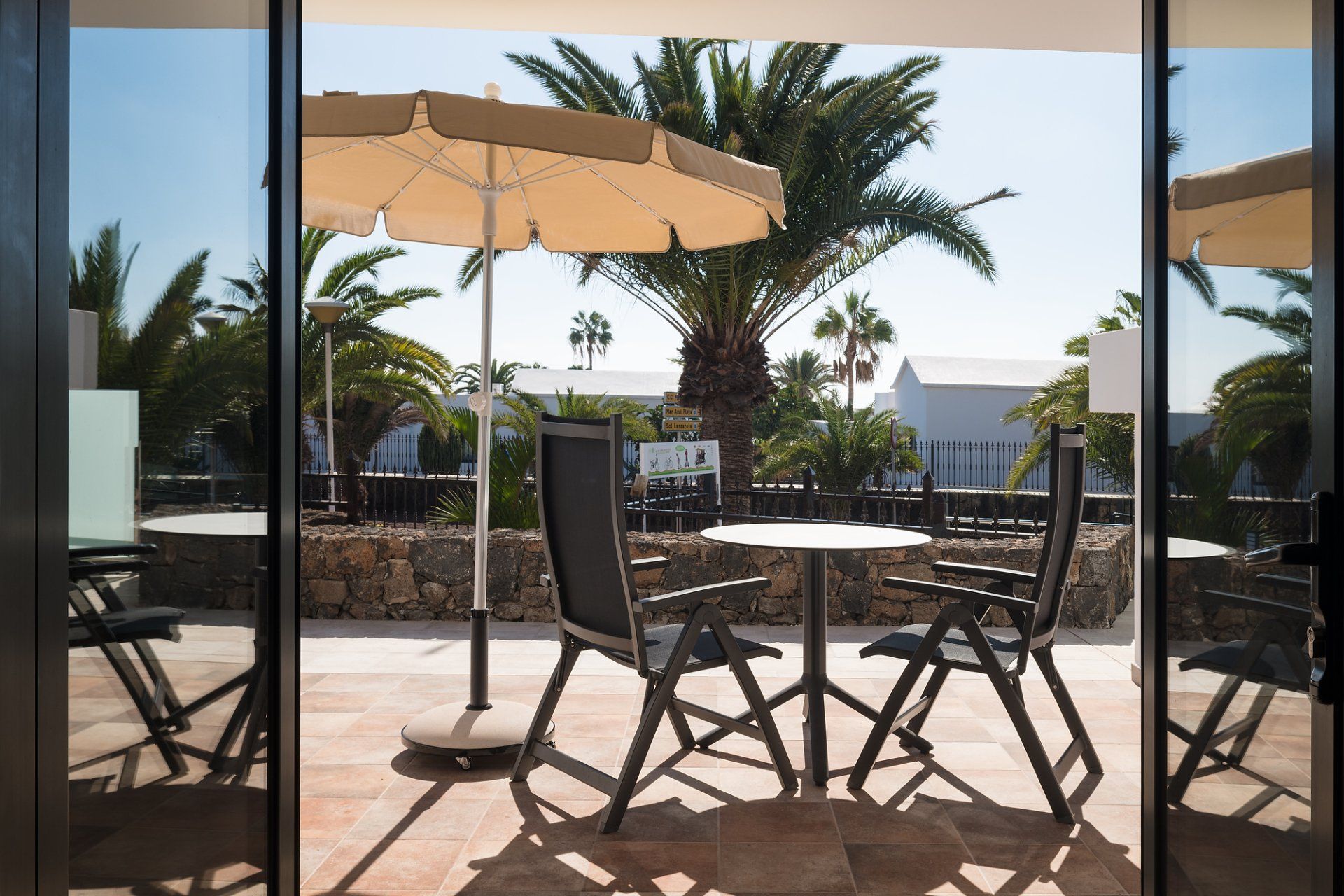 Patio con mesa, sillas, sombrilla y palmeras vistas a través de puertas corredizas de vidrio.