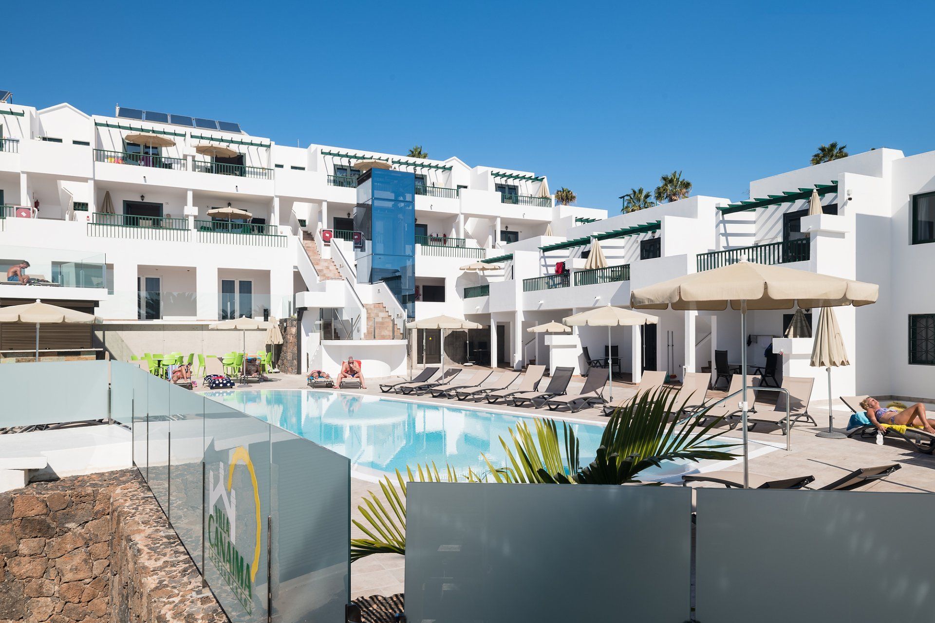 Área de piscina con edificios blancos, tumbonas y cielo azul.