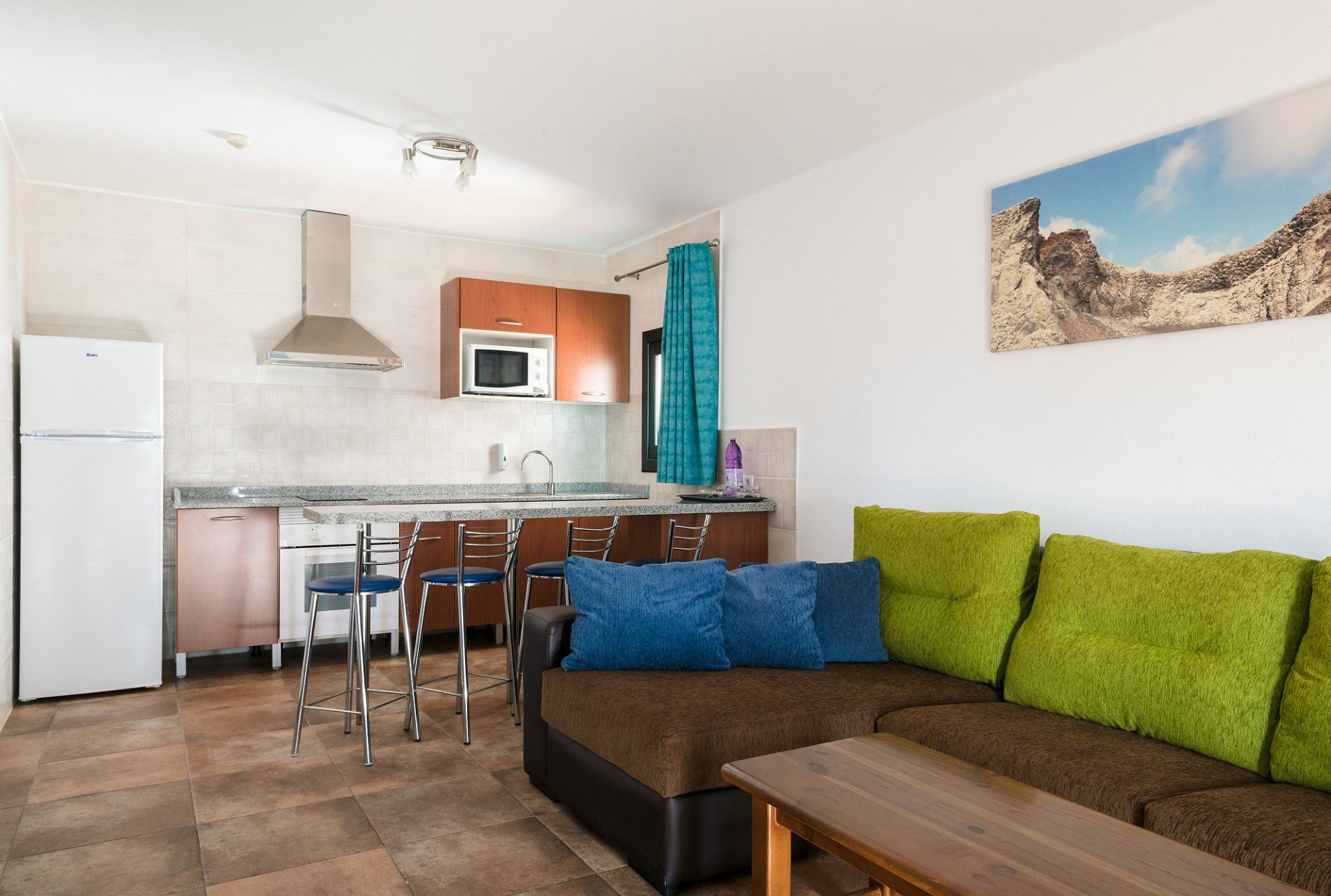 Pequeño apartamento tipo estudio con cocina, zona de estar y un sofá marrón con almohadas verdes y azules.