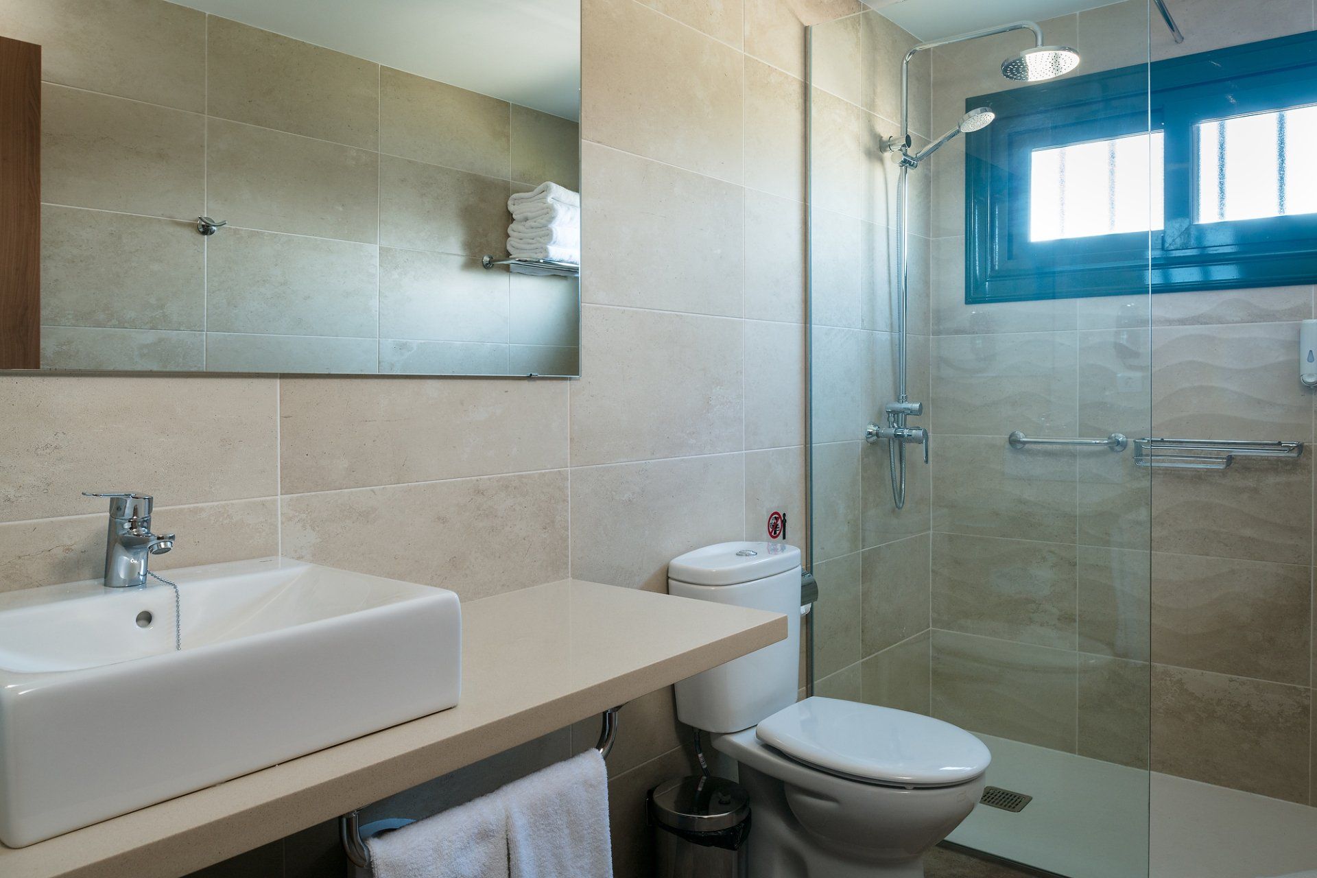 Baño con lavabo rectangular, inodoro, ducha de cristal, azulejos beige y pequeña ventana.