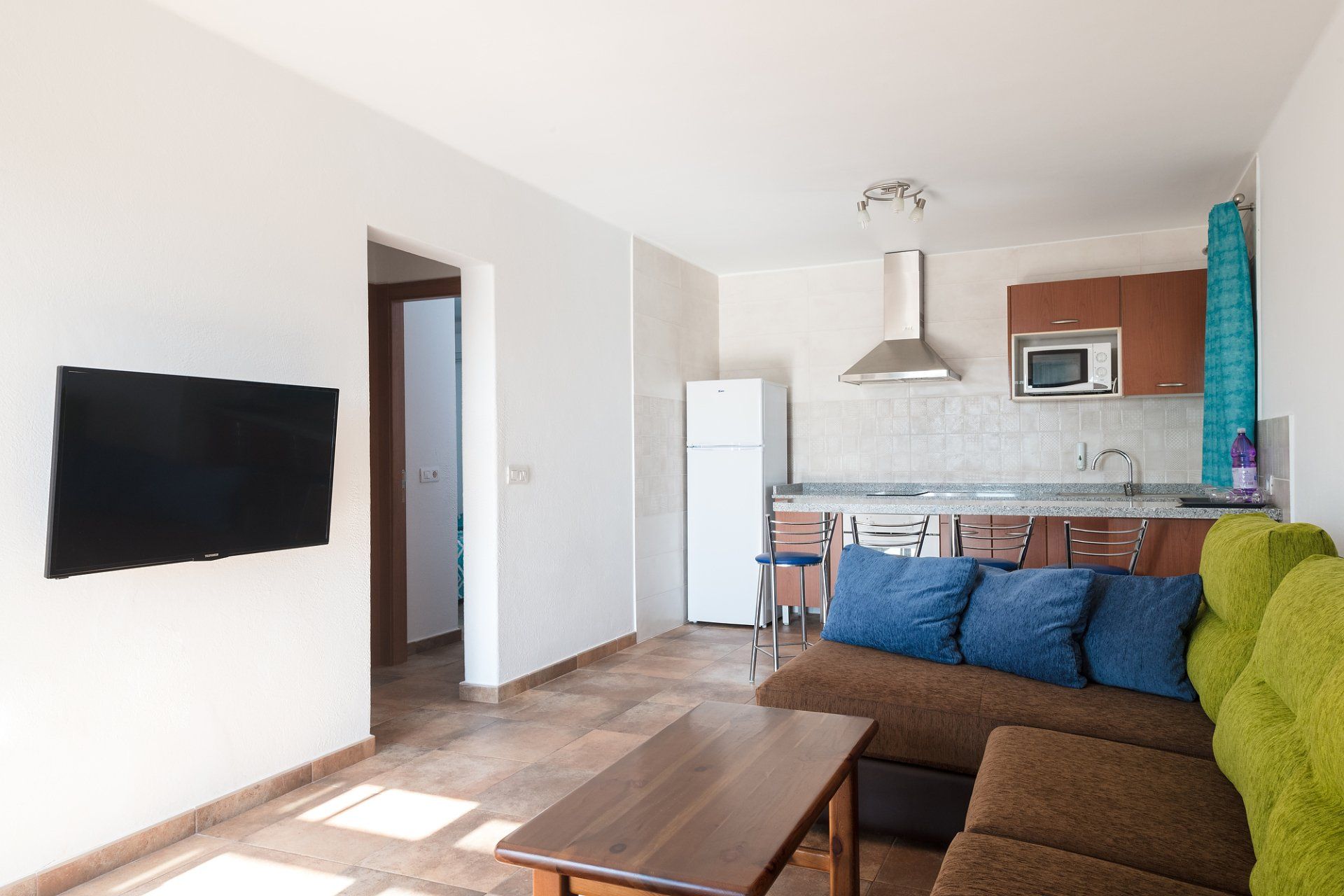Sala de estar con cocina: Sofá marrón, TV en la pared, área de cocina con refrigerador y microondas.