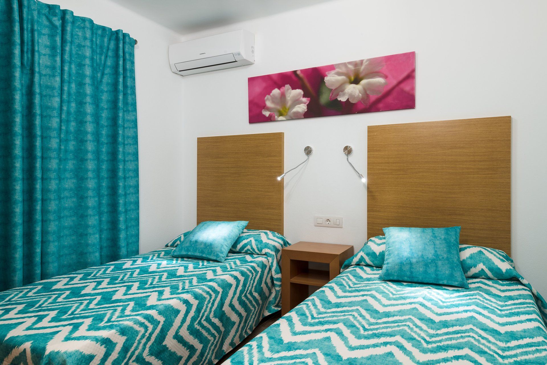 Dos camas con mantas turquesas de espiga, habitación luminosa con cortinas y cuadro floral.