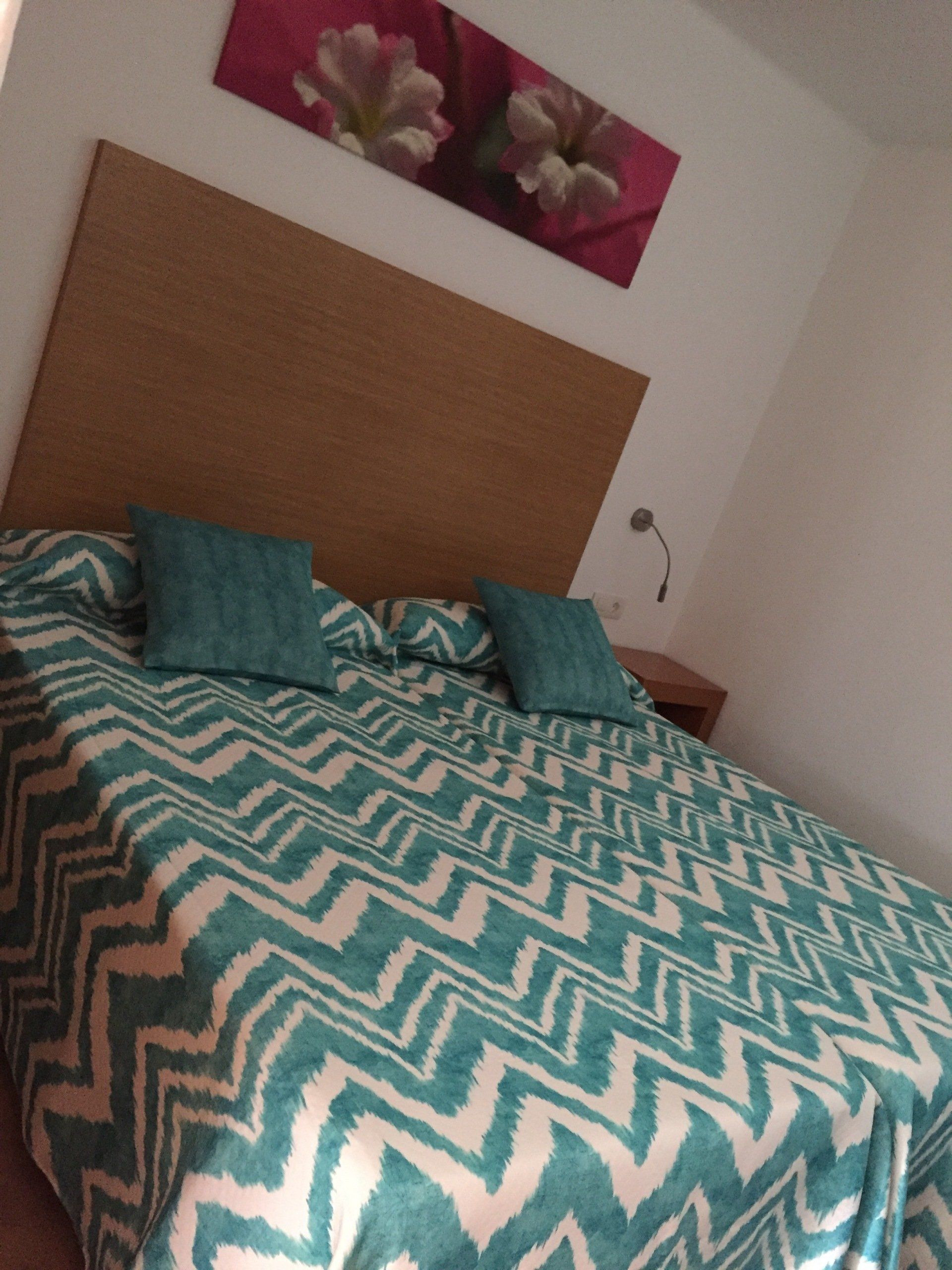 Dormitorio con colcha con estampado en zigzag en tonos turquesa y blanco, almohadas en tonos turquesa y cabecero de madera.