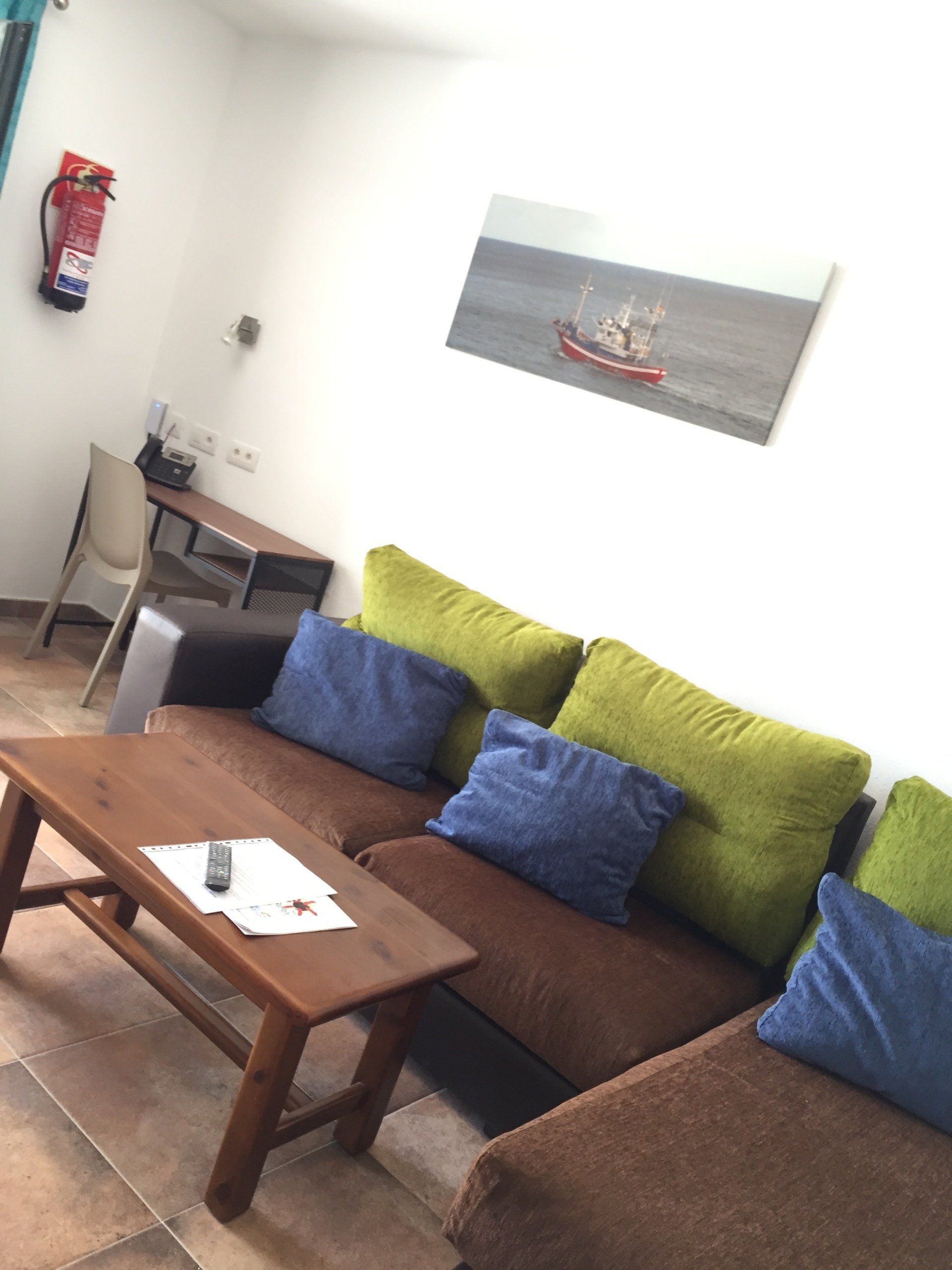 Sala de estar con sofá marrón, mesa de madera, almohadas verdes y azules y un cuadro.