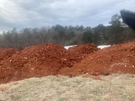 Piles of reddish mulch or soil on grass with trees and cloudy sky in the background