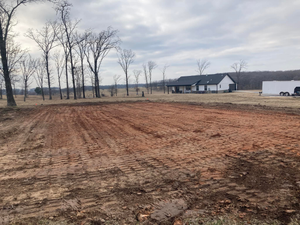 Cleared dirt lot with bare trees and a white house in the distance under a cloudy sky