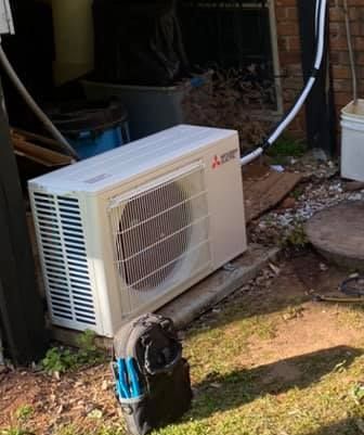 A Mitsubishi outdoor air conditioning unit sits on a concrete pad near a brick wall with a tool bag in the foreground.